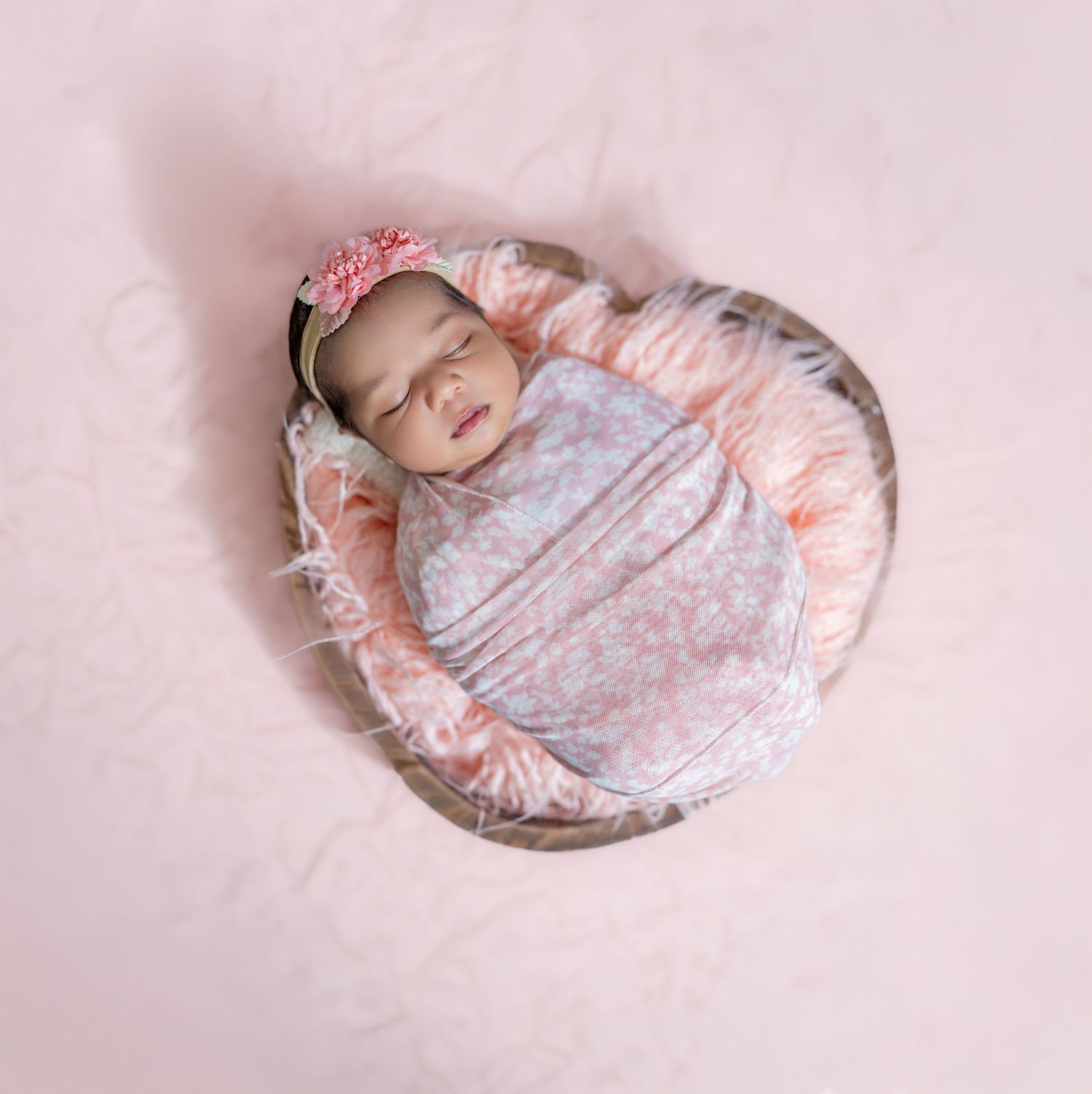 Newborn baby girl swaddled in floral wrap with pink headband, sleeping peacefully in a rustic bowl on pink fur.