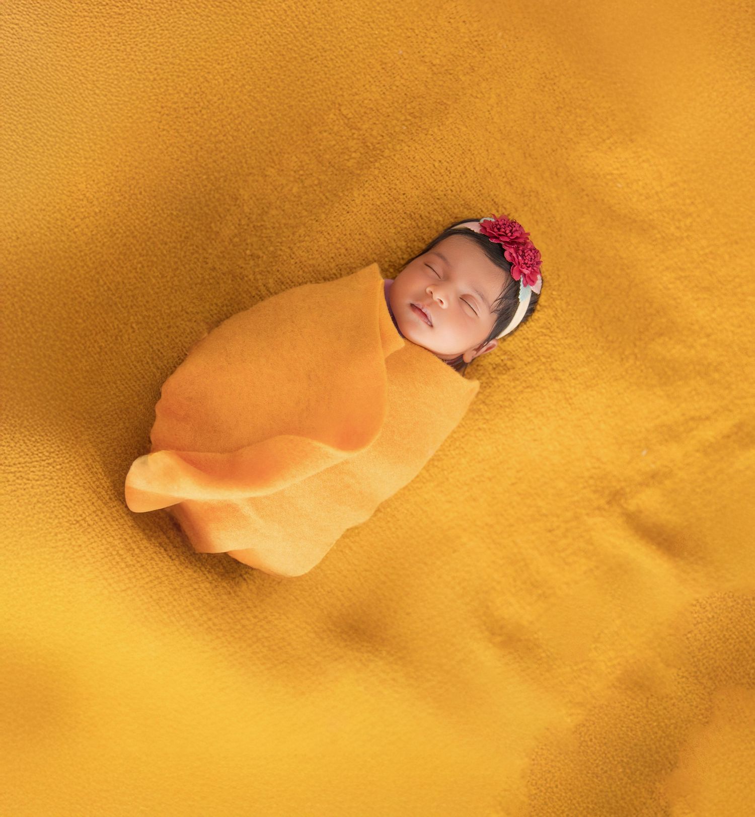 Newborn baby wrapped in soft orange swaddle blanket resting on golden yellow background with pink headband.
