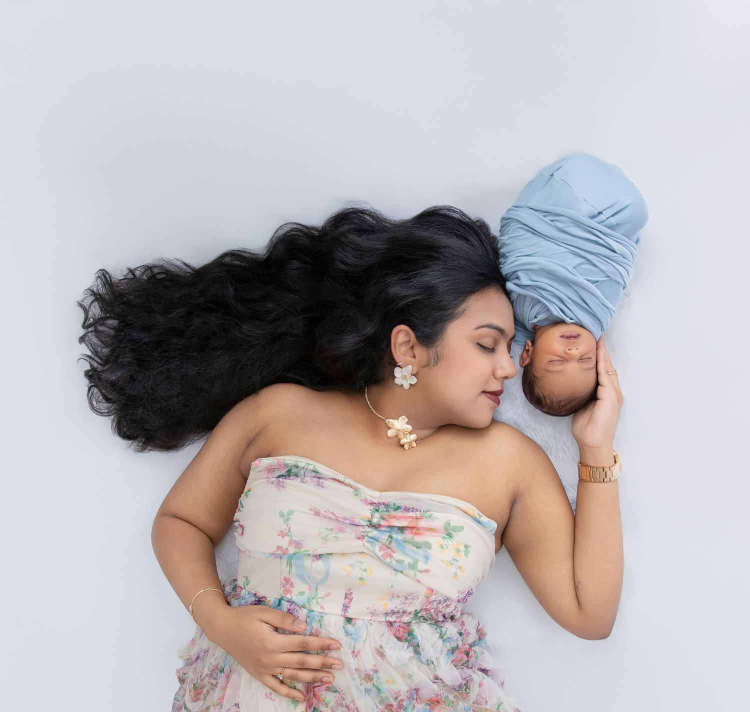 A woman in a floral dress lies peacefully on a white surface with long flowing black hair and a baby in a blue hat.