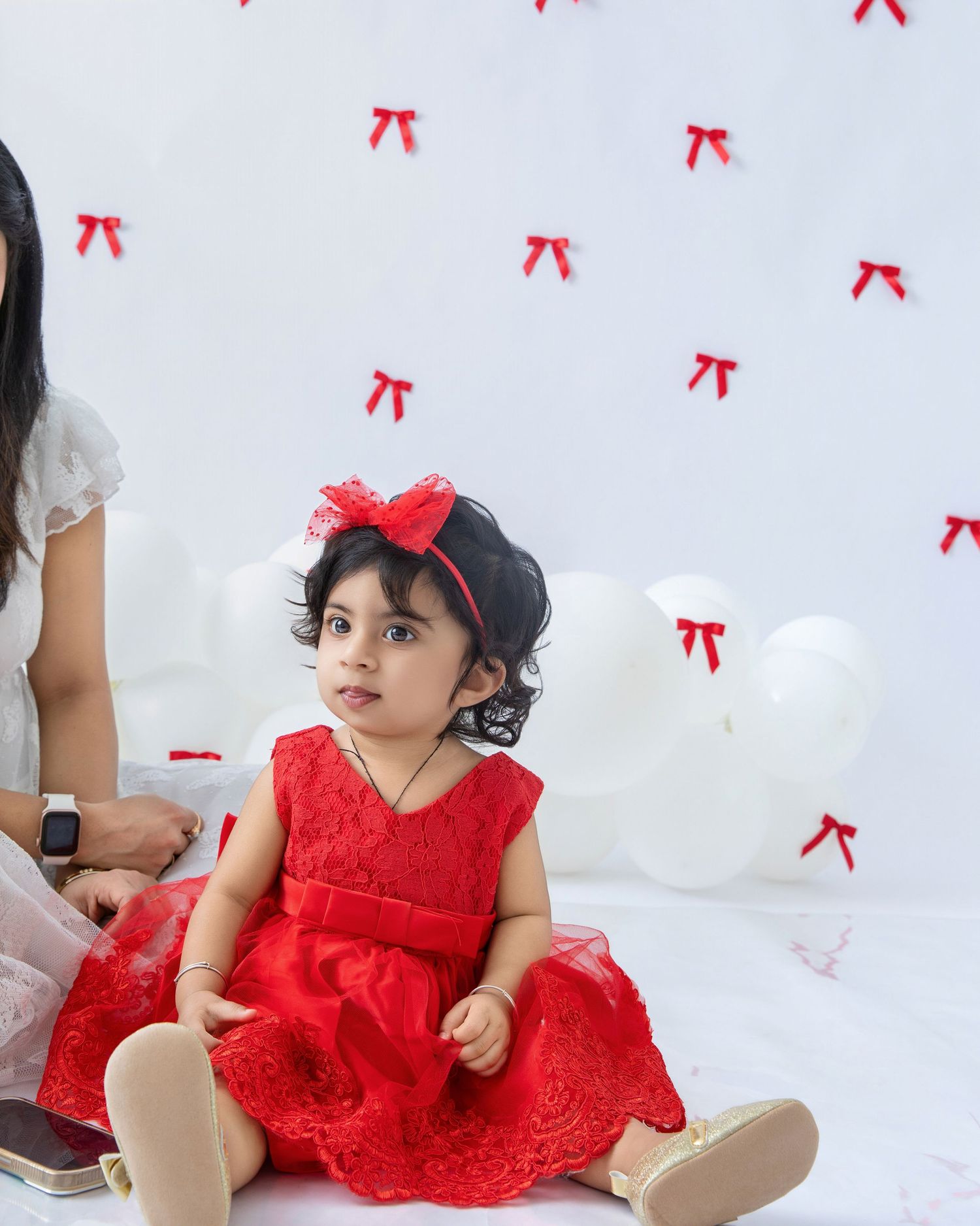 A baby in a red lace dress and bow sits on a white background with scattered red bow decorations.