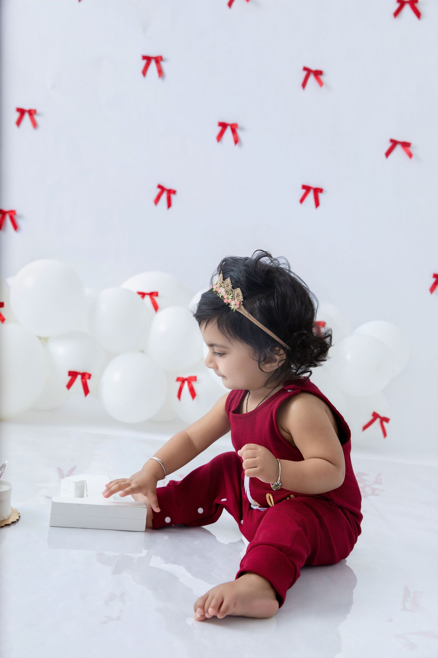 Baby in red velvet dress crawls on white floor with red bows decorating the background.