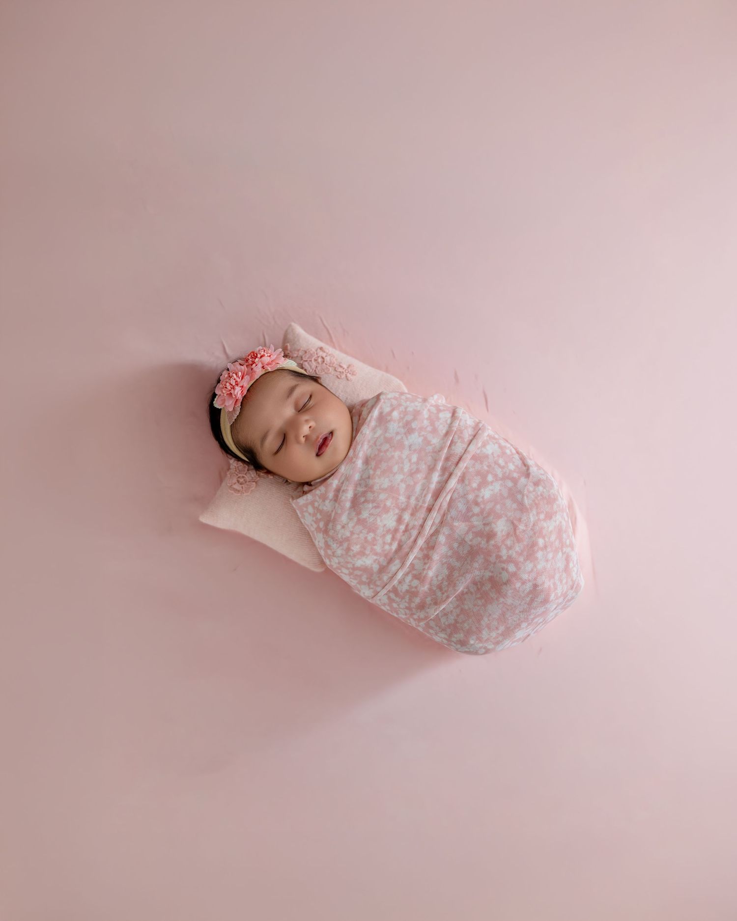 Sleeping newborn baby swaddled in pink floral wrap with pink flower headband on soft pink background.