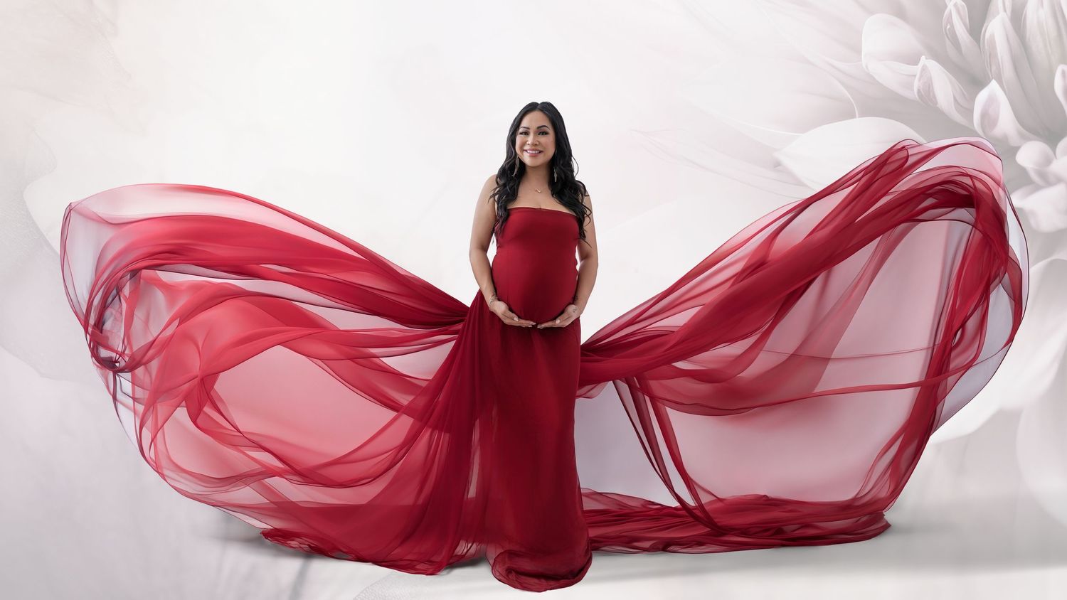 Pregnant woman in flowing red maternity gown with dramatic fabric wings against white floral background.