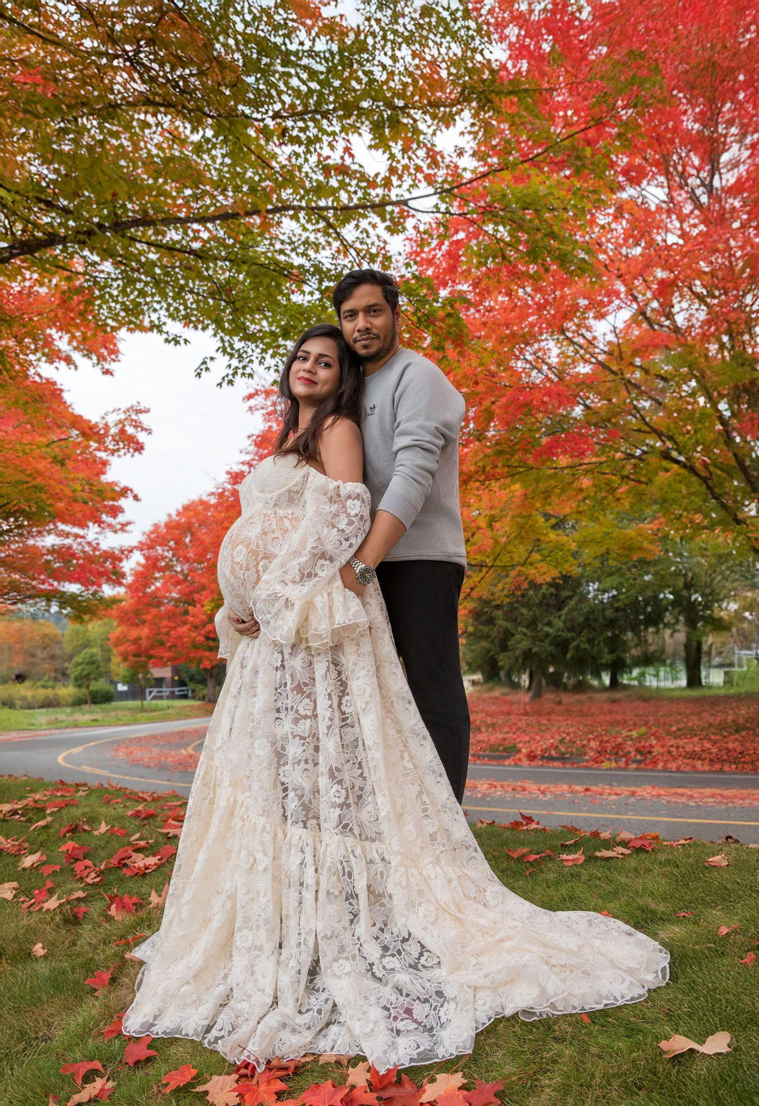 Pregnant woman in white lace gown with man in gray sweater amid stunning red autumn maple trees in a park.