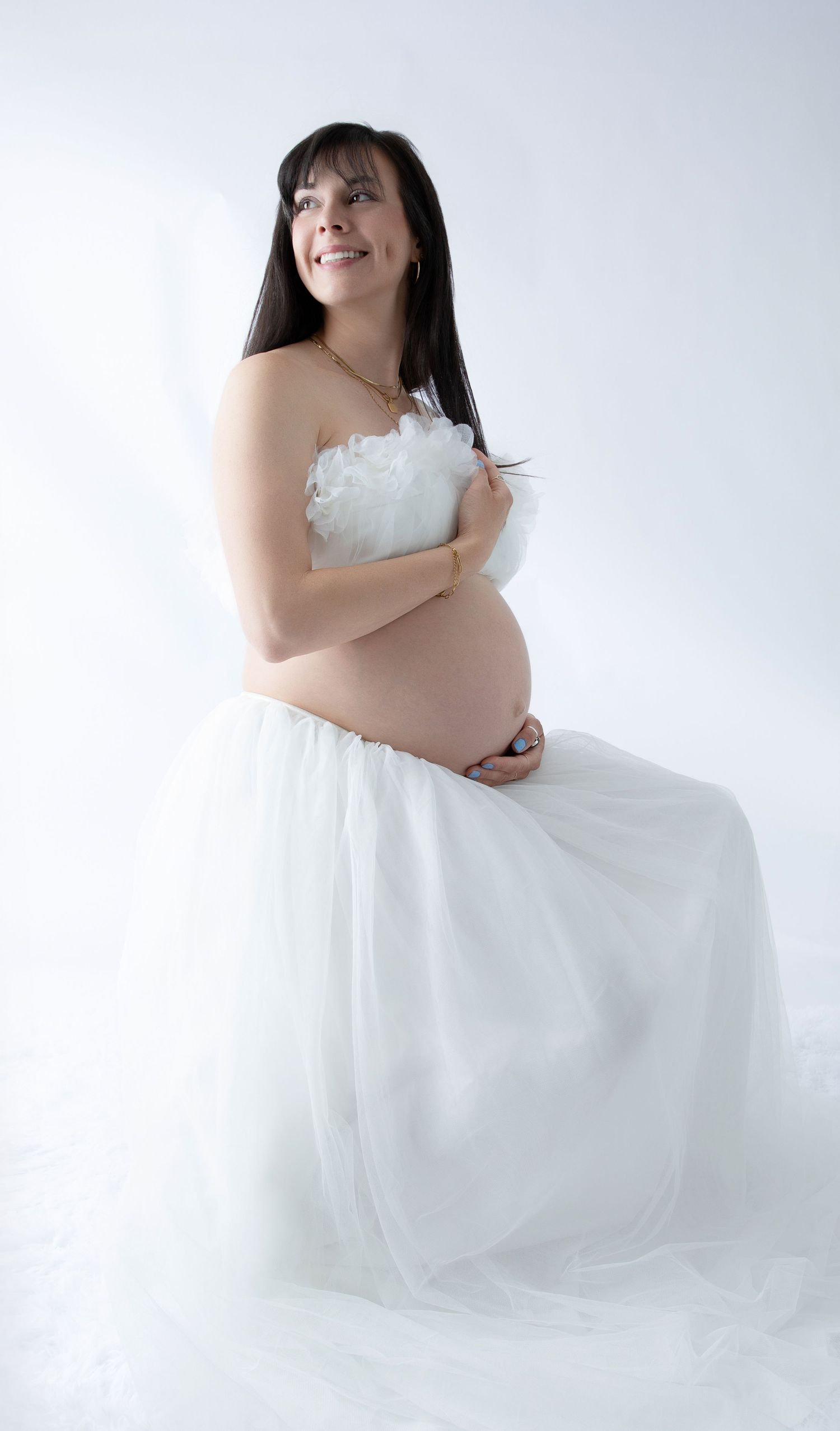 Smiling pregnant woman in white strapless tulle gown cradling her baby bump during a maternity photo shoot.