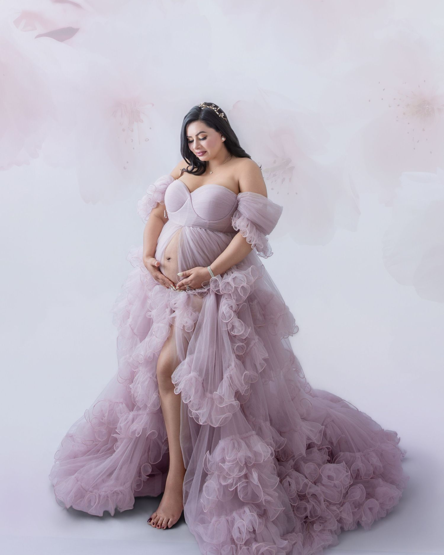 Pregnant woman in elegant dusty pink ruffled tulle maternity gown posing gracefully in a soft floral studio setting.