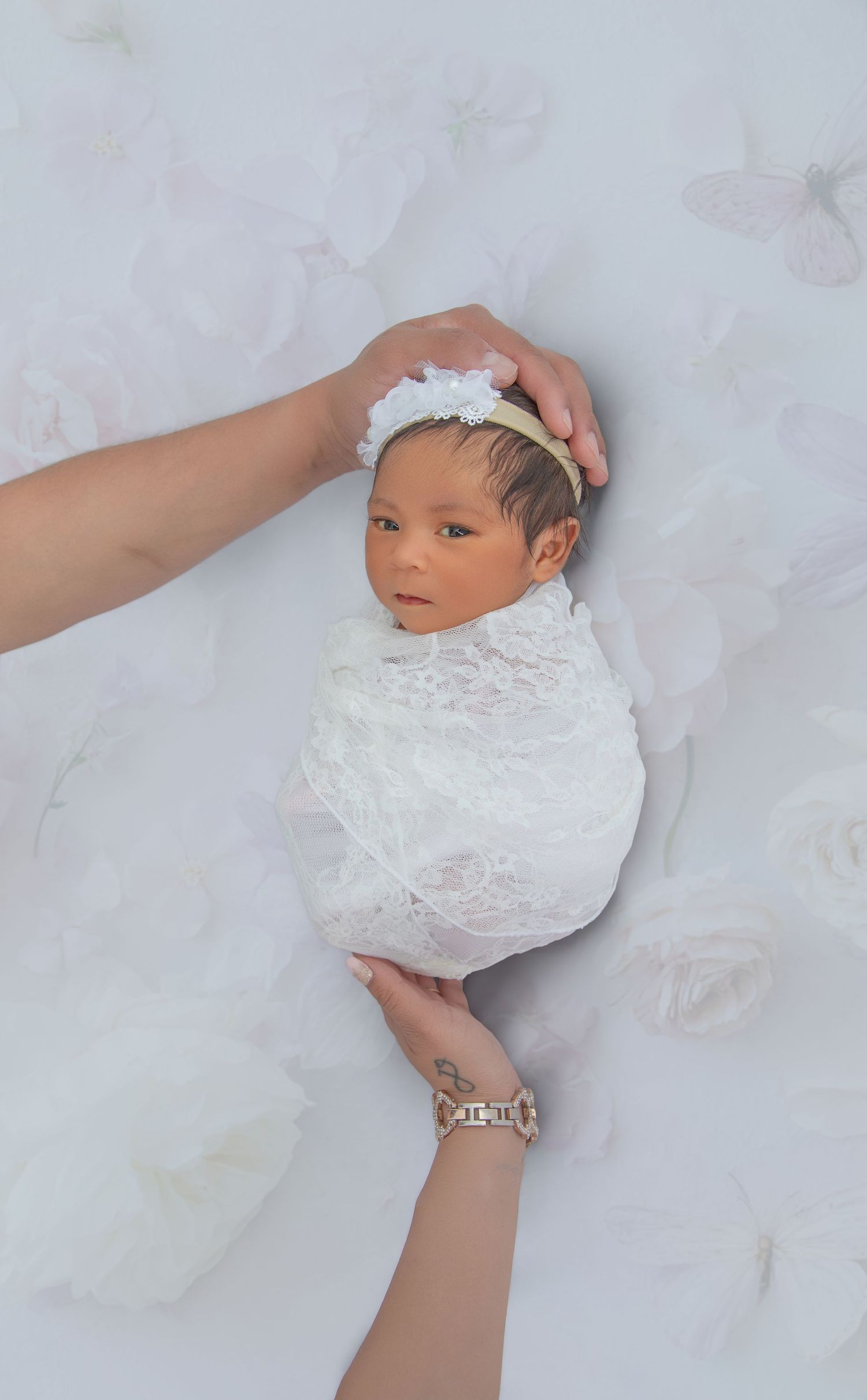 Parents hands gently holding swaddled newborn baby girl in white lace wrap with floral headband.