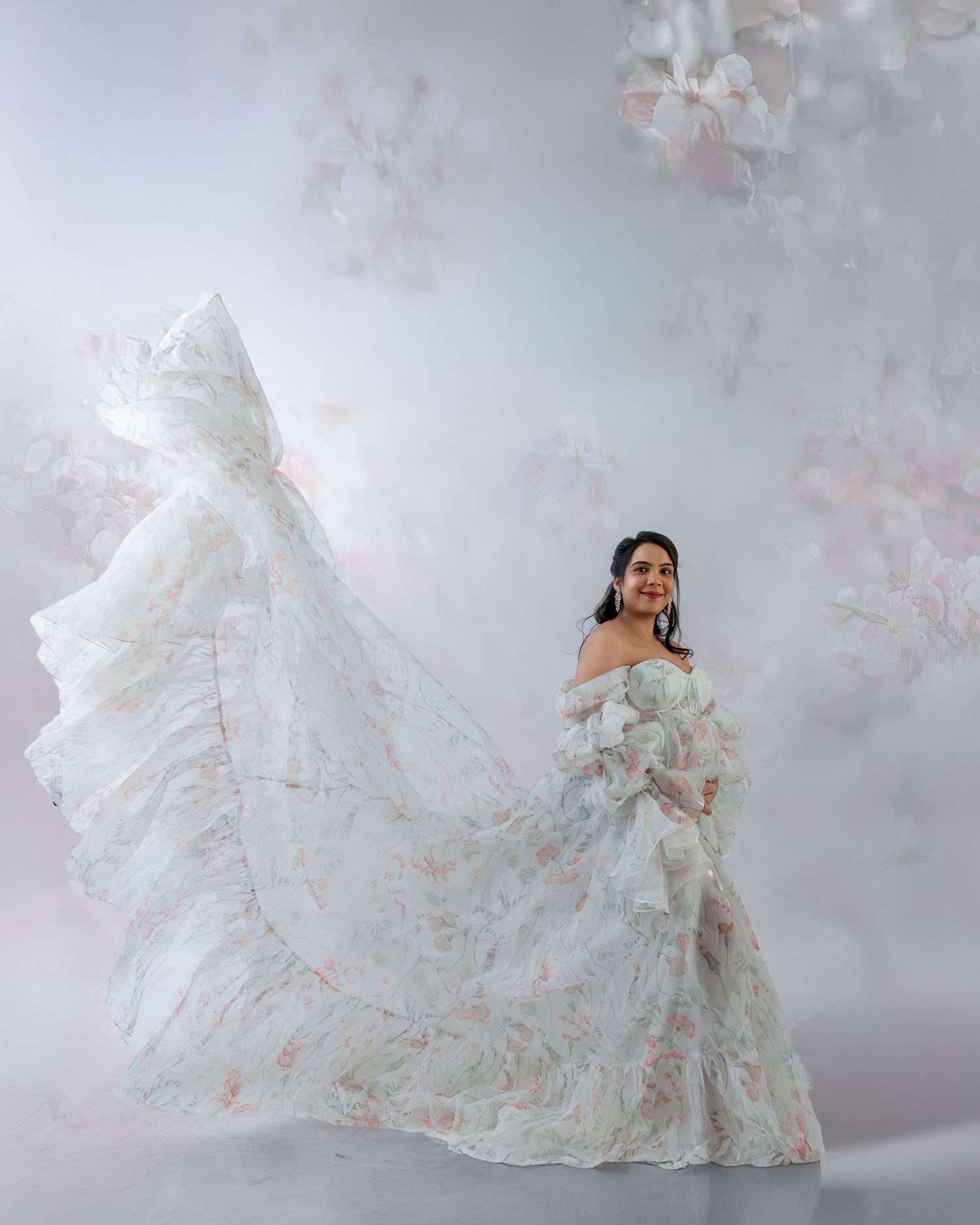 Woman in flowing white floral off-shoulder gown with dramatic train, posing elegantly against soft floral backdrop.