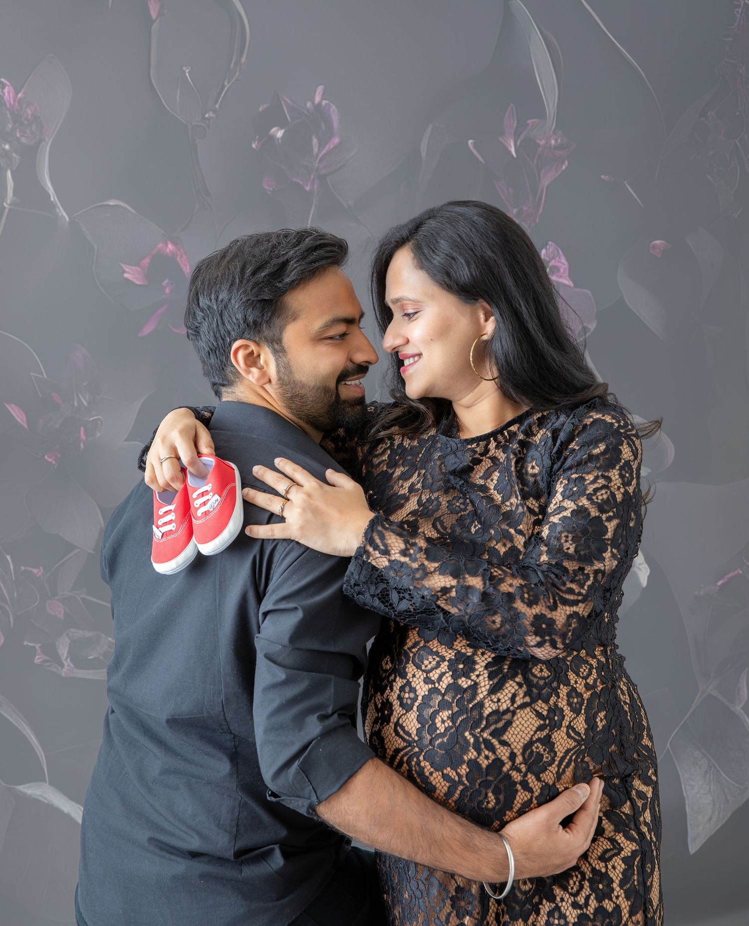 Happy expecting couple embracing and holding tiny red baby shoes during maternity photo session.