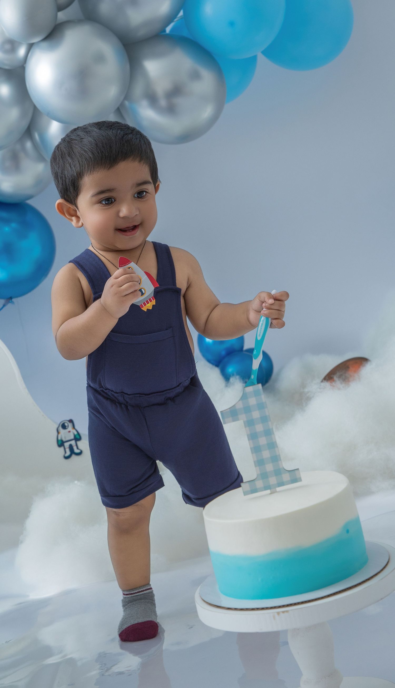 A cake smash photo session with blue and silver balloons featuring an ombre blue cake and navy overalls.