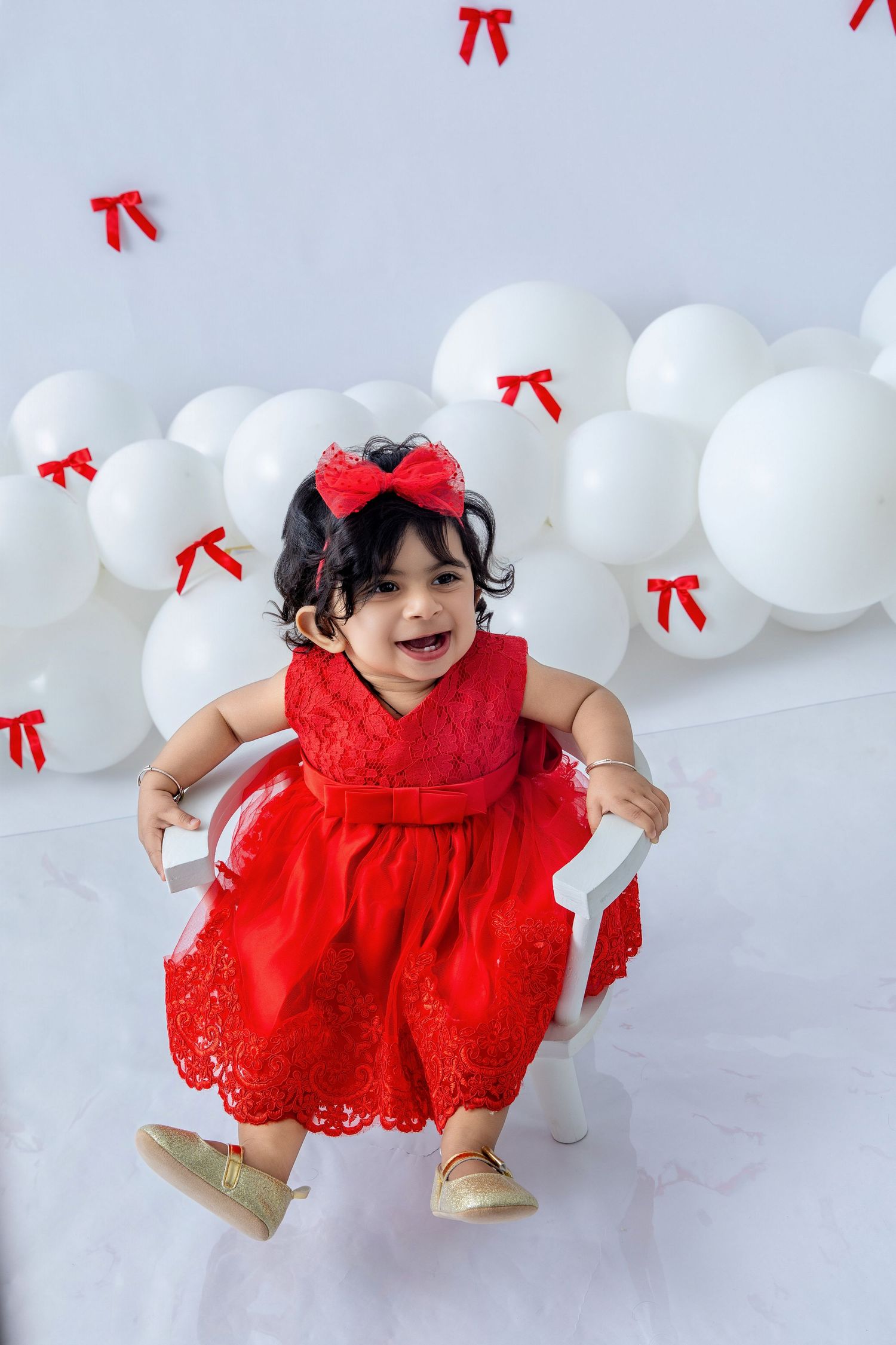 Playful baby crawling on white floor with red bows and balloons in background.