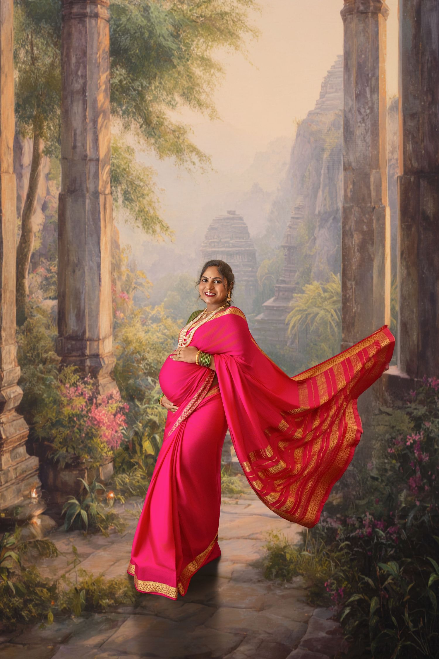 Pregnant woman in vibrant pink saree with gold border poses gracefully before scenic temple backdrop with lush greenery.