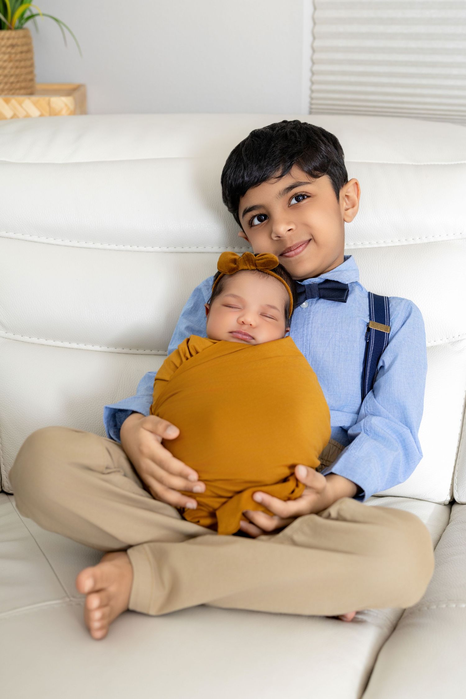 Young boy in blue shirt and suspenders sits cross-legged on white sofa holding sleeping swaddled newborn sibling.