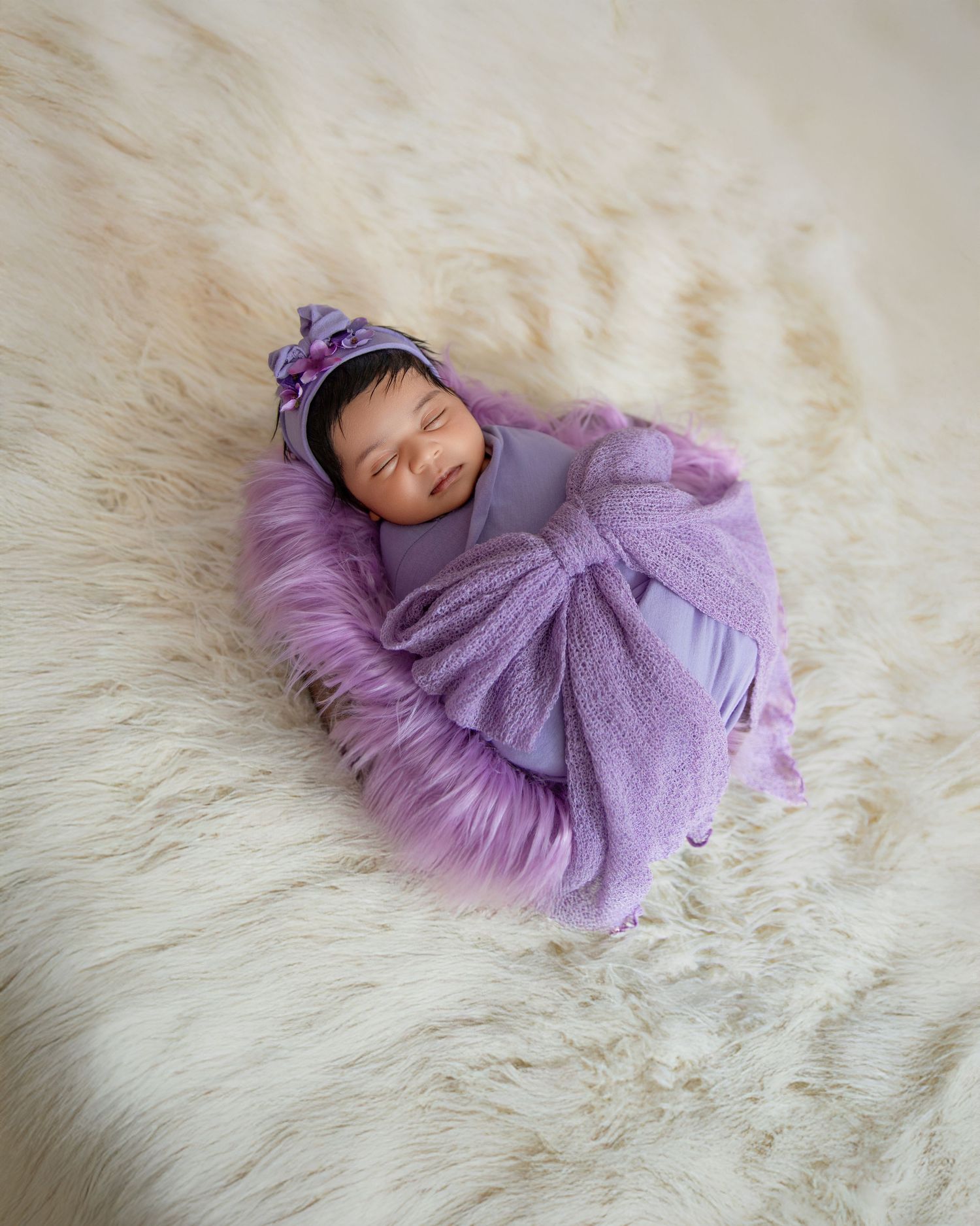 A sleeping newborn in a soft purple knit wrap lies on a plush cream-colored fur blanket.