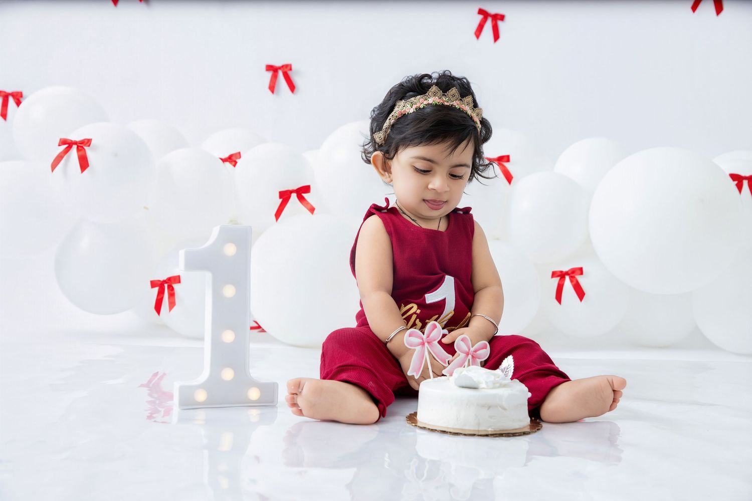 Baby in red outfit examines white birthday cake during photo session.