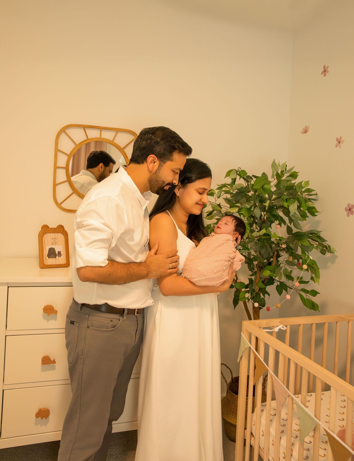 A couple in white clothing embraces their newborn baby in a nursery decorated with a wooden crib and houseplant.