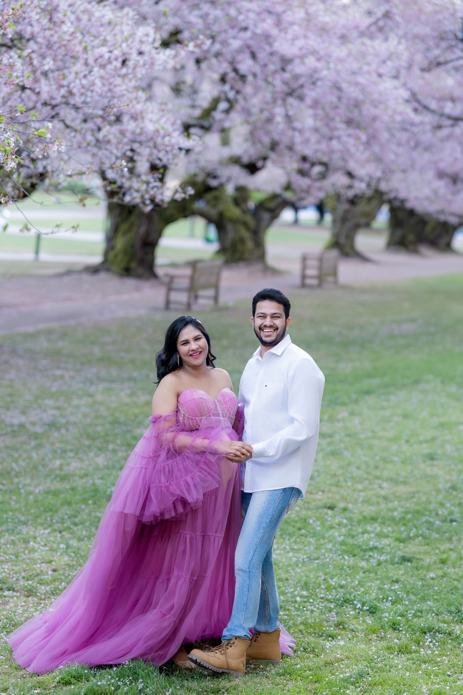 Couple posing under blooming cherry blossom trees, woman in purple tulle gown, man in white shirt and jeans.