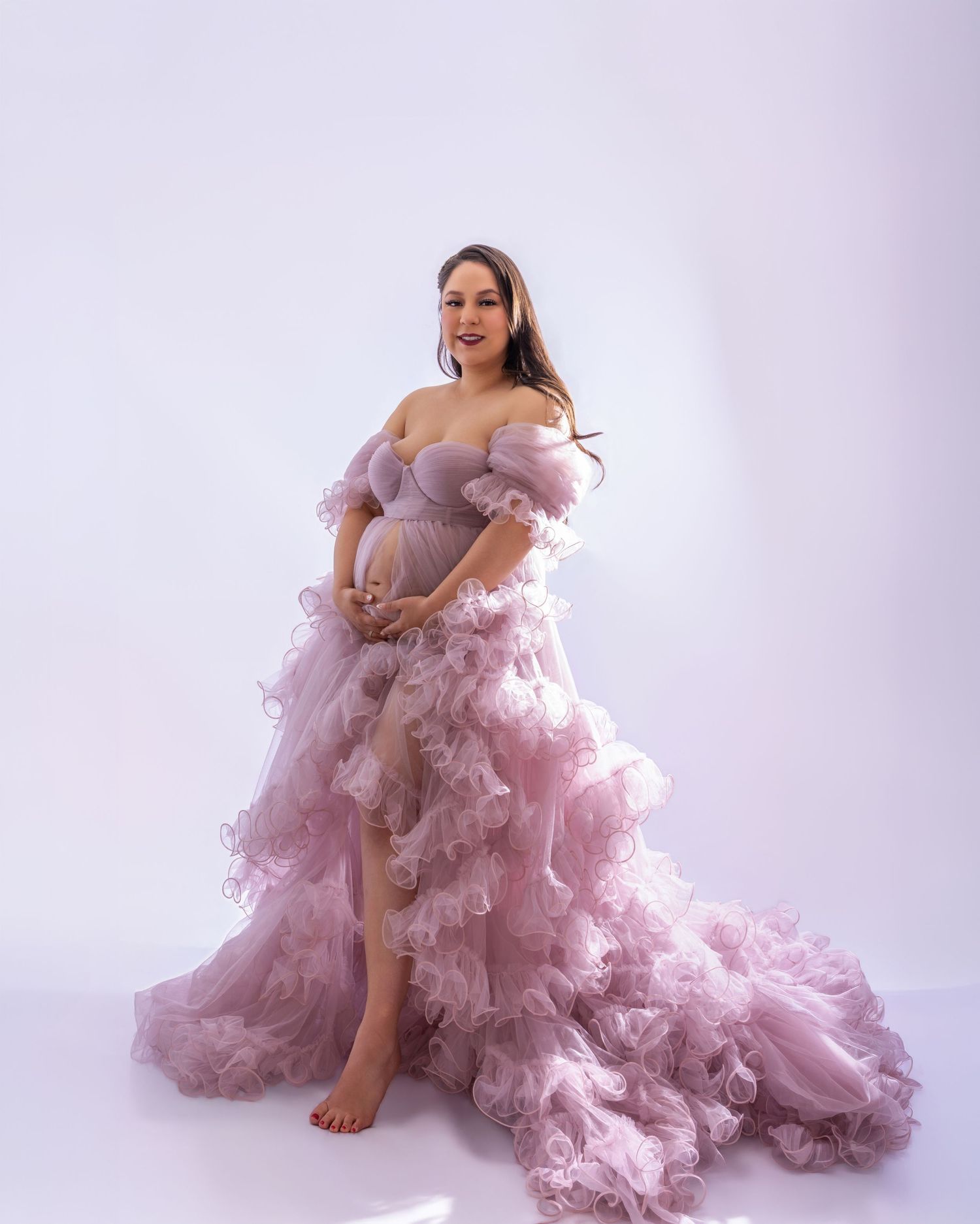 Pregnant woman in a flowing pink ruffled off-shoulder tulle maternity gown posing gracefully in a studio.