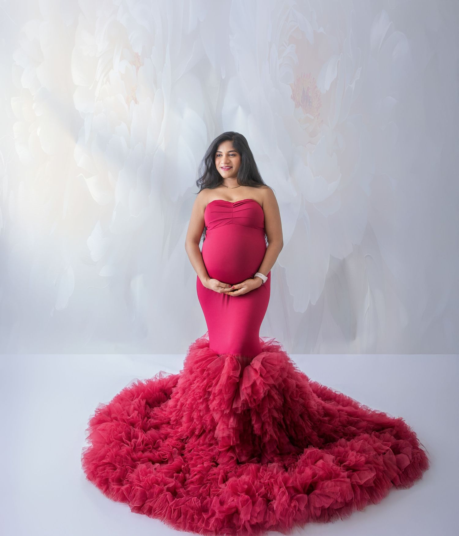 Pregnant woman in elegant strapless red mermaid gown with ruffled train poses gracefully in a studio maternity photoshoot.