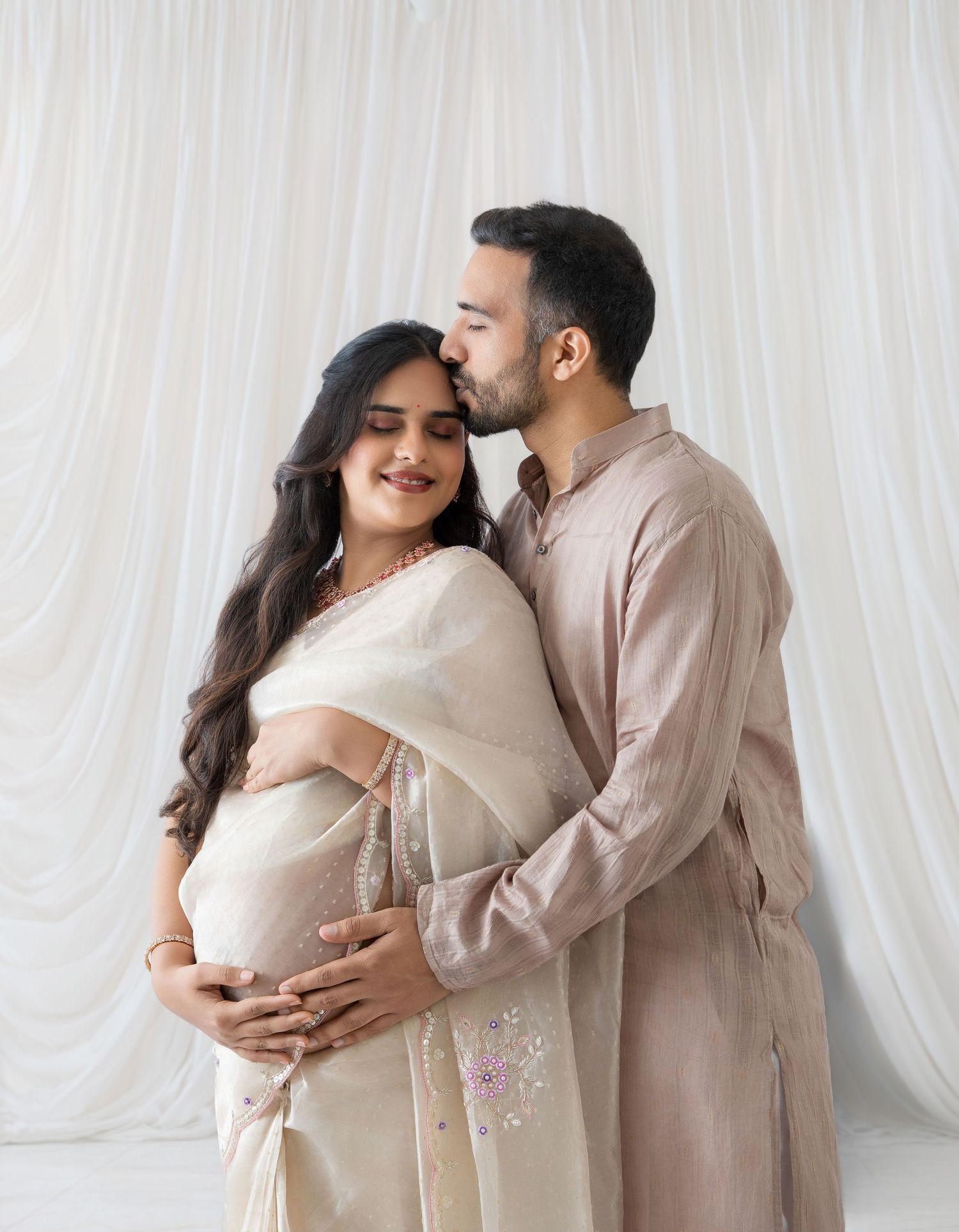 A couple in traditional Indian attire shares a tender maternity moment, the man kissing his pregnant wife's head.