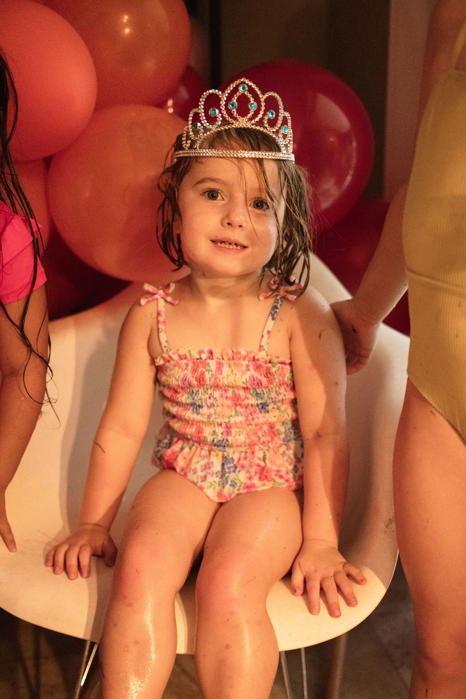 A small child in a floral swimsuit sits surrounded by red balloons and party decorations.