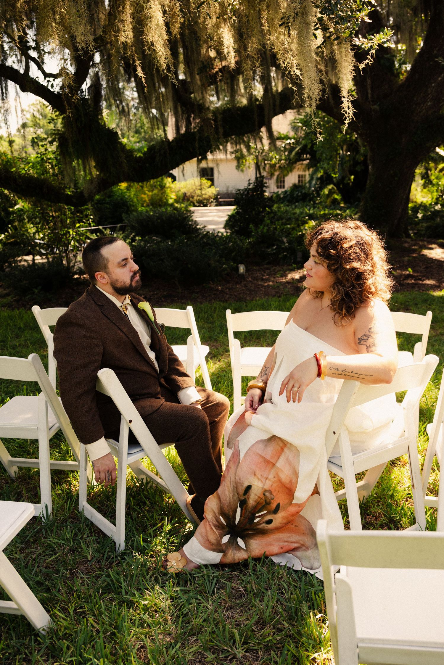 A couple sits on white chairs under Spanish moss at an outdoor wedding venue.
