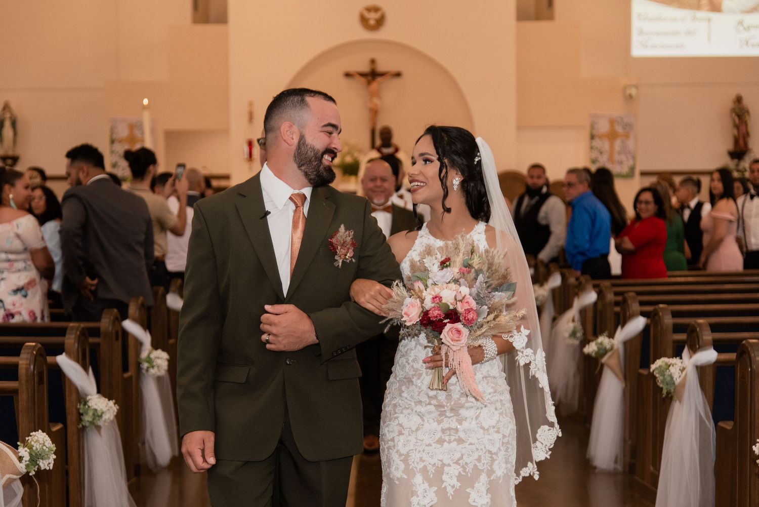 A couple walks down the church aisle after their wedding ceremony surrounded by decorated pews.
