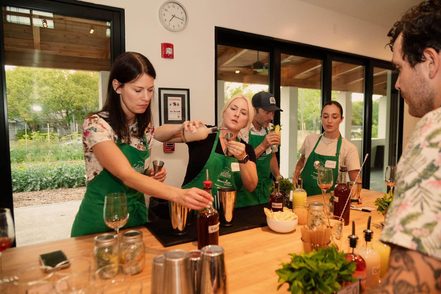People in green aprons learning bartending skills in a bright modern kitchen space.