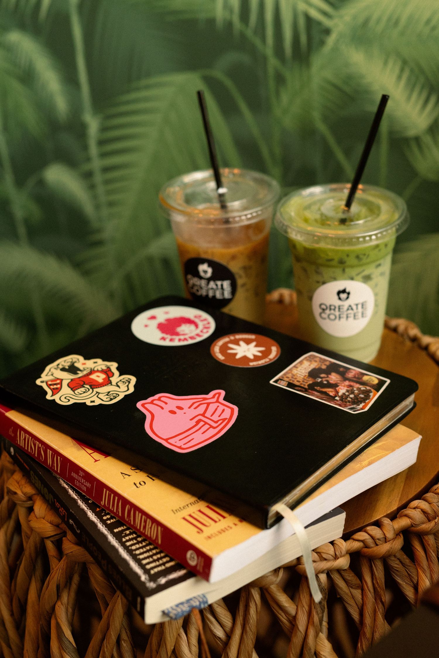 Books with decorative stickers and two iced drinks on woven surface against tropical leaf backdrop.