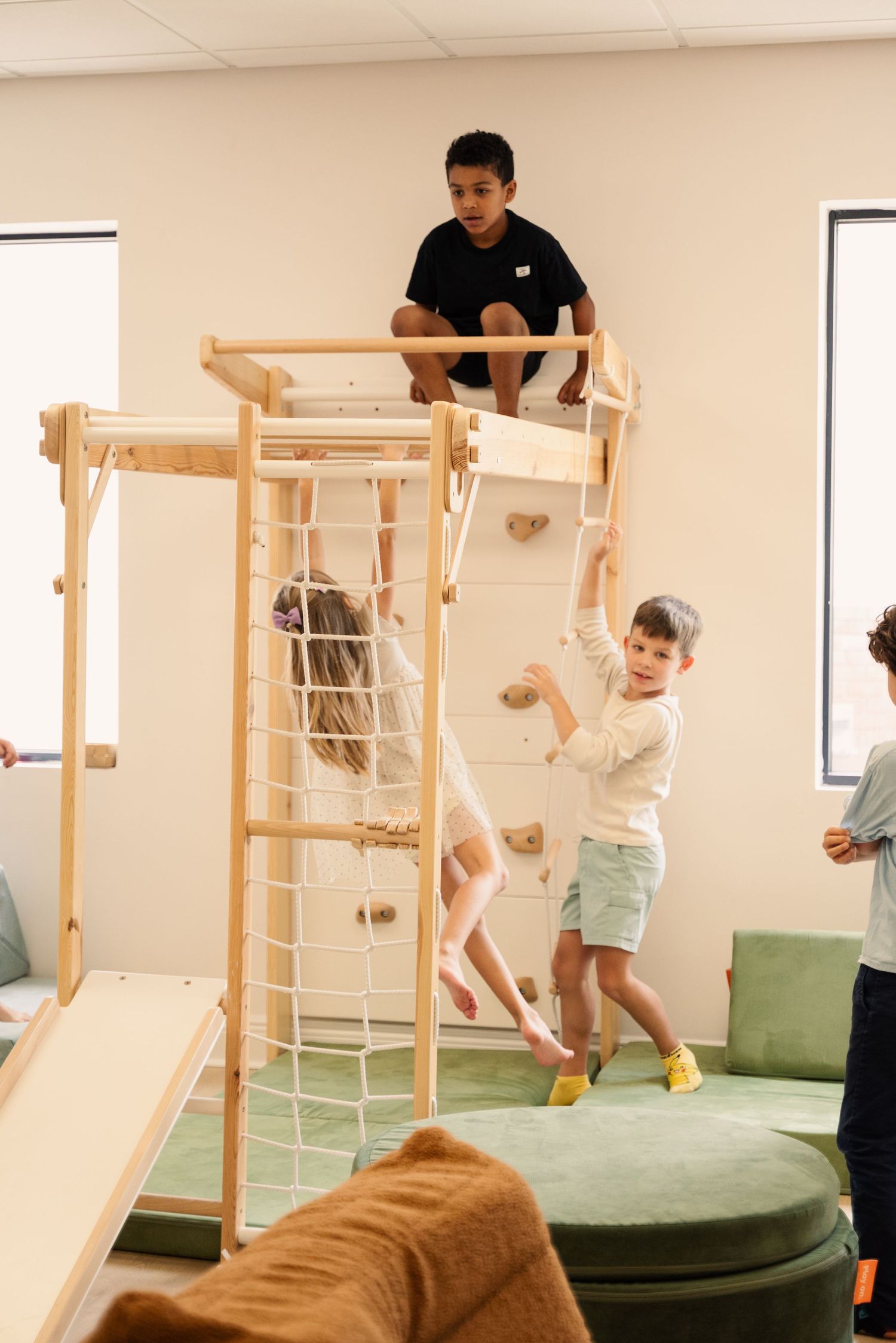 Children playing on wooden climbing equipment and exercise structures in an indoor playroom.