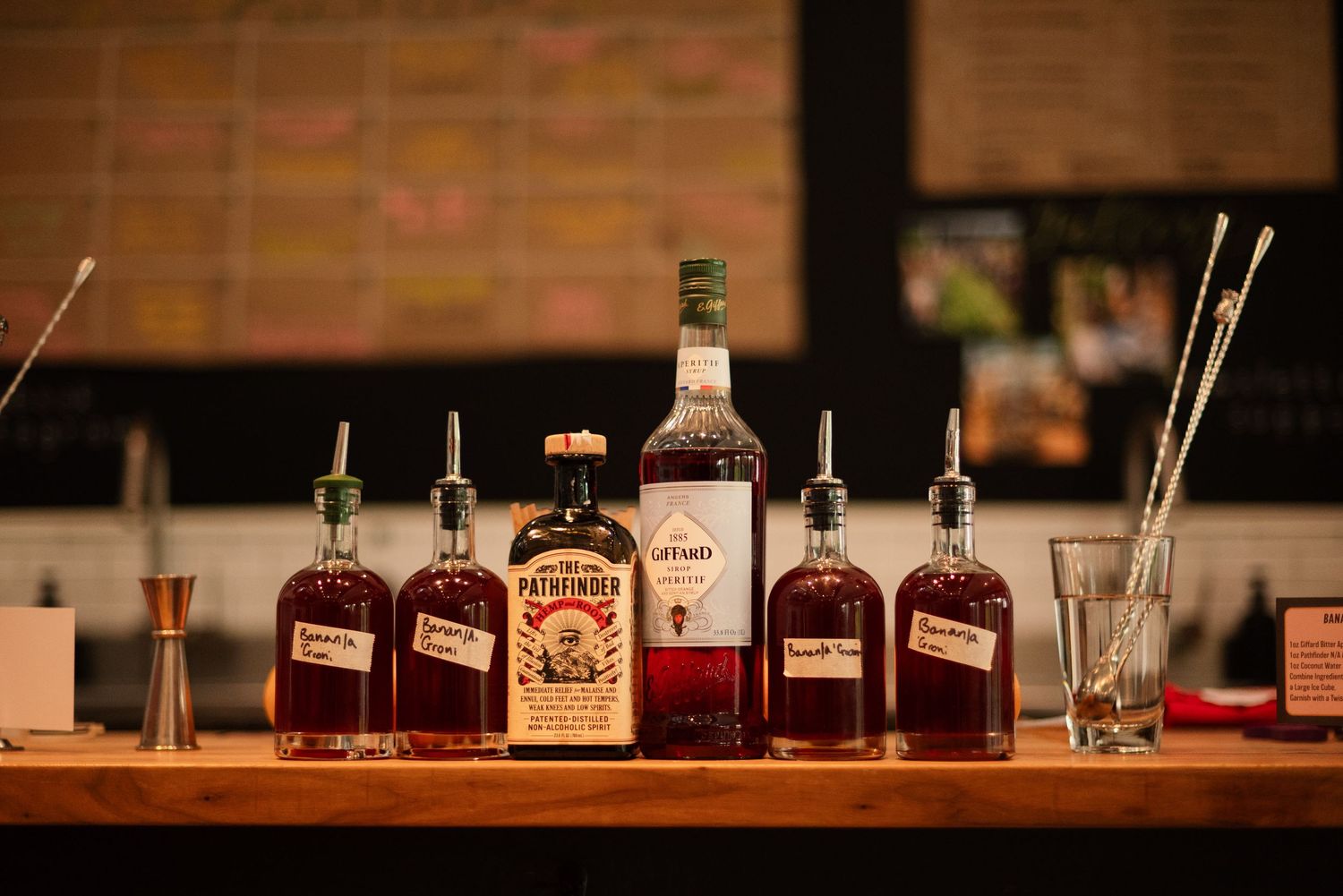 Row of whiskey bottles arranged on wooden bar counter with pour spouts and mixing tools.