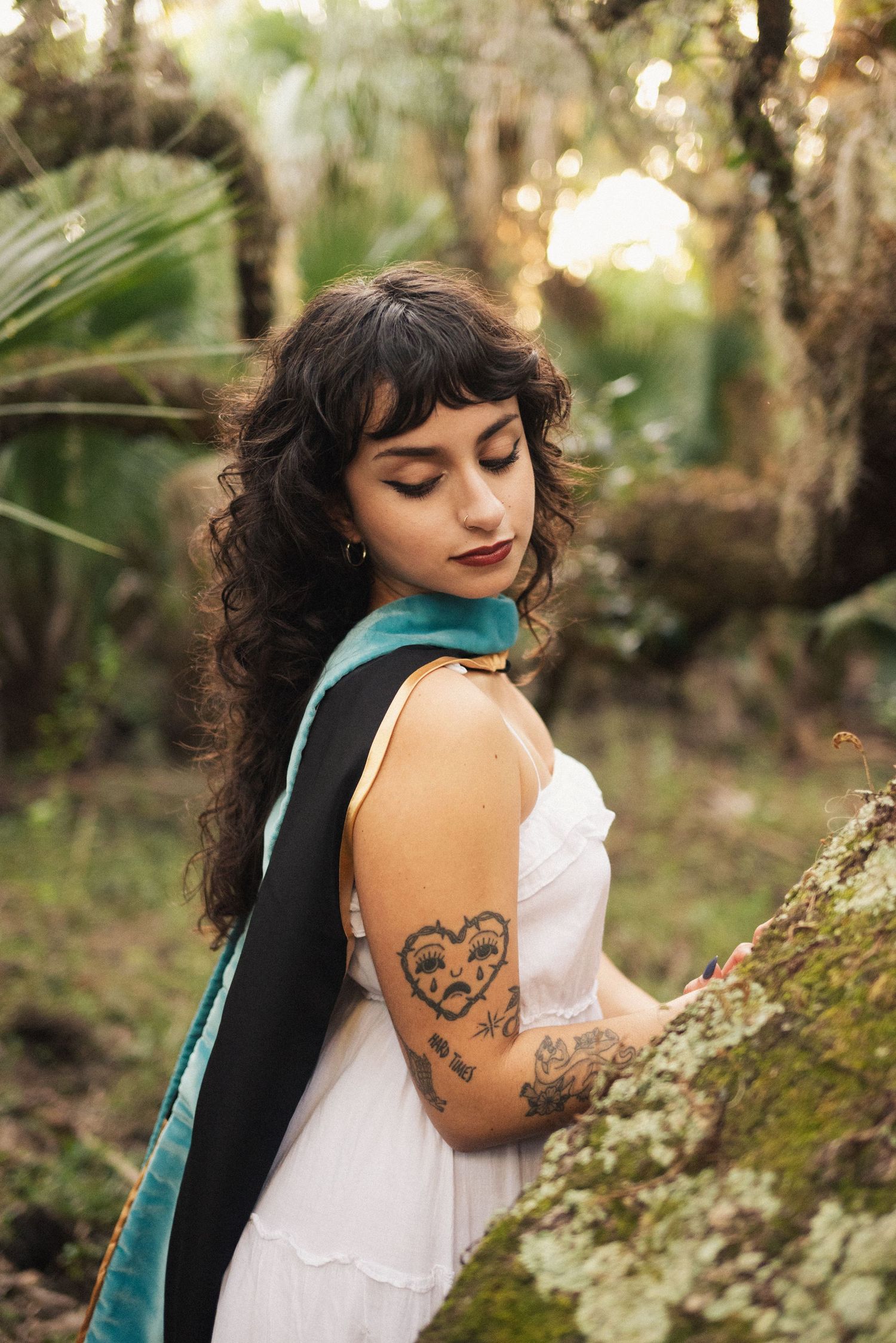 Person in white dress with tattoos poses against mossy trees during golden hour photography session.
