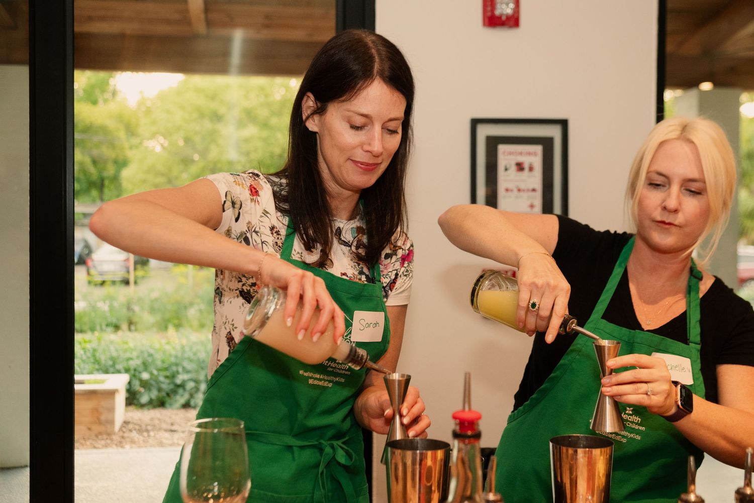 Students in green aprons practicing cocktail making techniques at a wooden bar counter.