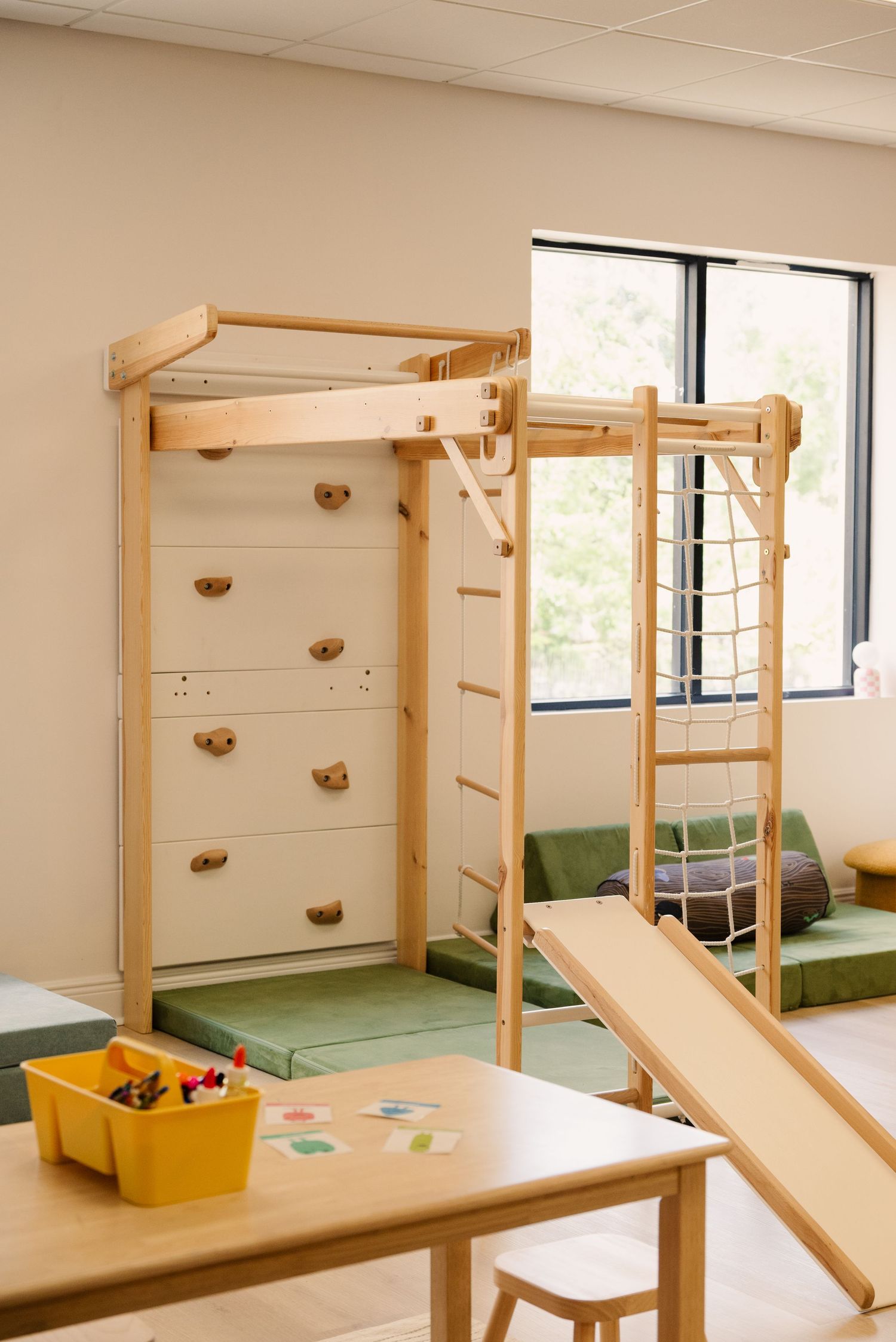 Indoor play area with wooden climbing frames and green mats installed along windows.