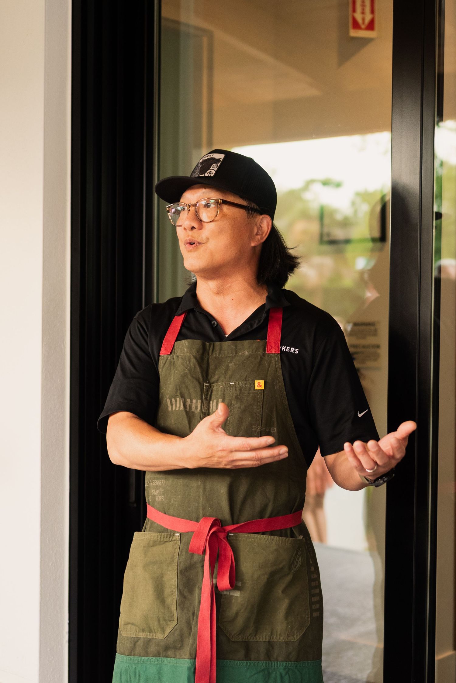 A restaurant worker in an apron and baseball cap gesturing while standing in a doorway.