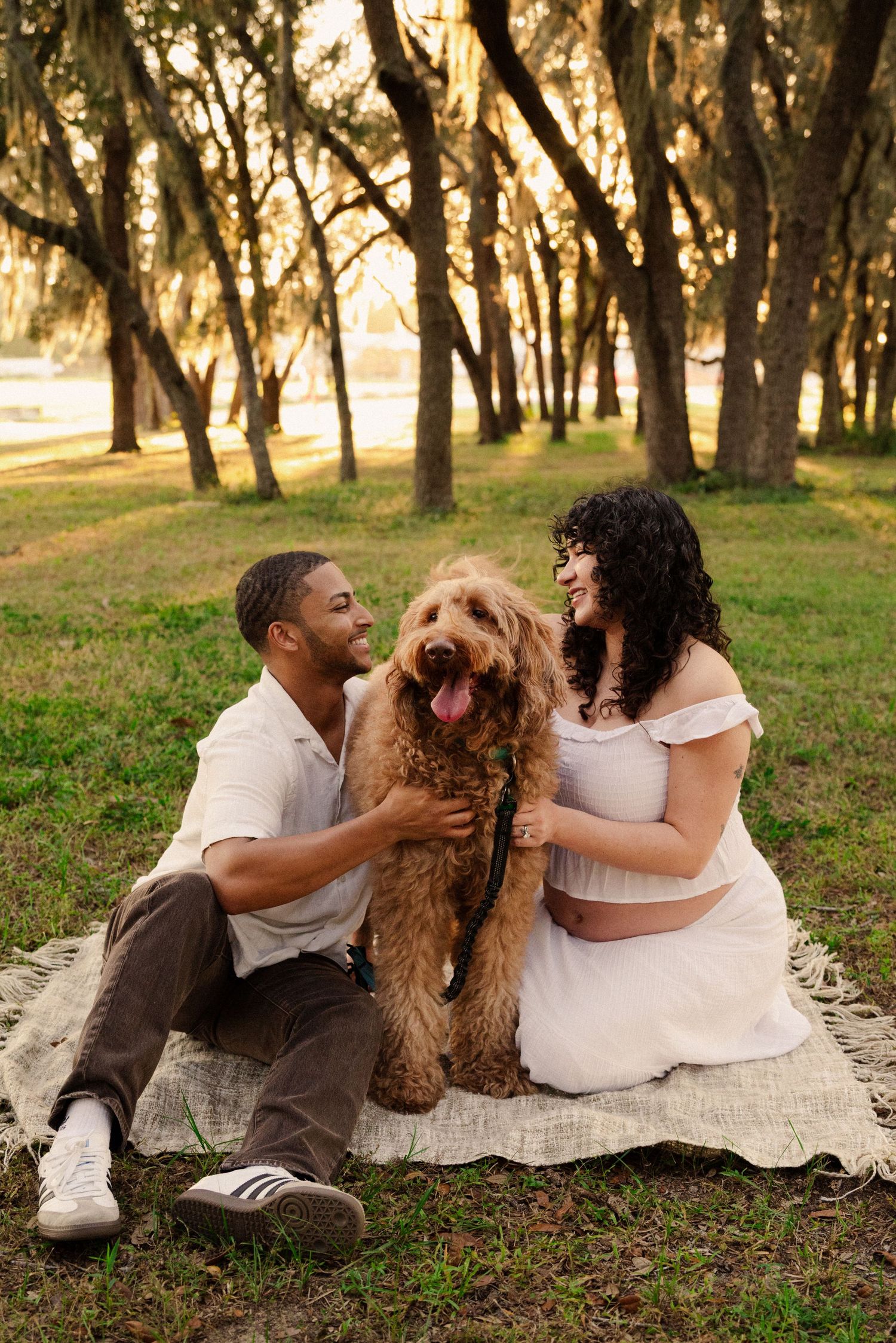 A couple relaxes with their fluffy brown dog on a picnic blanket in a wooded park.