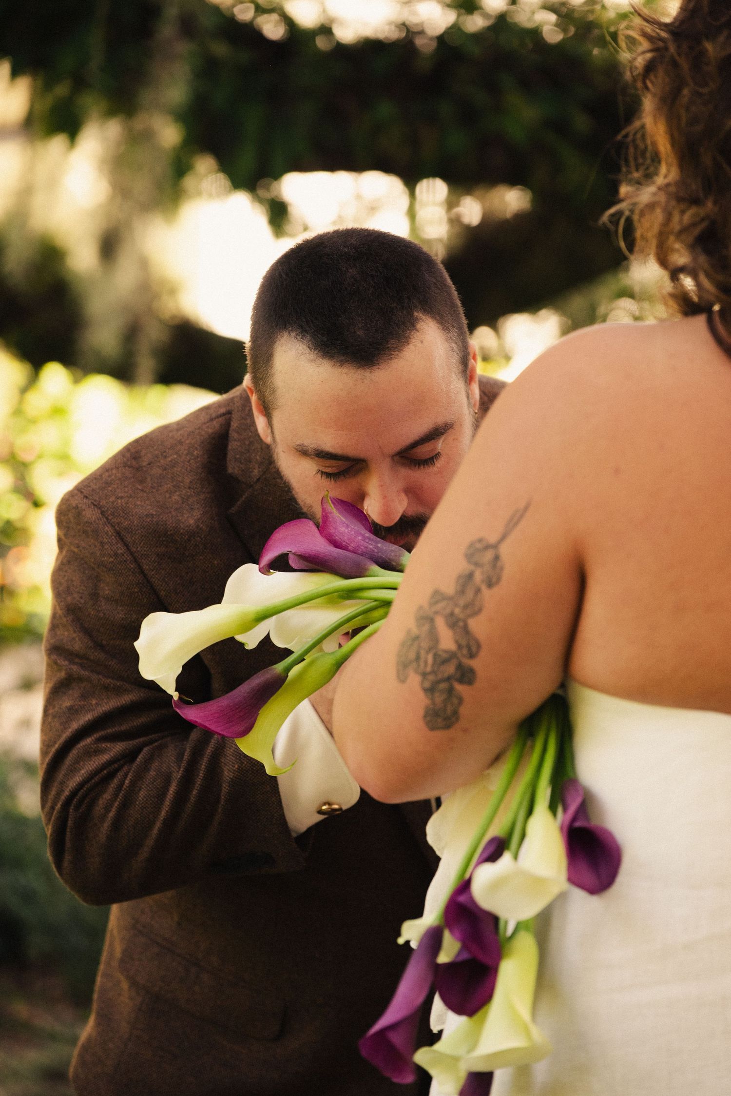 Person in brown suit smells bouquet of white and purple flowers against white dress.