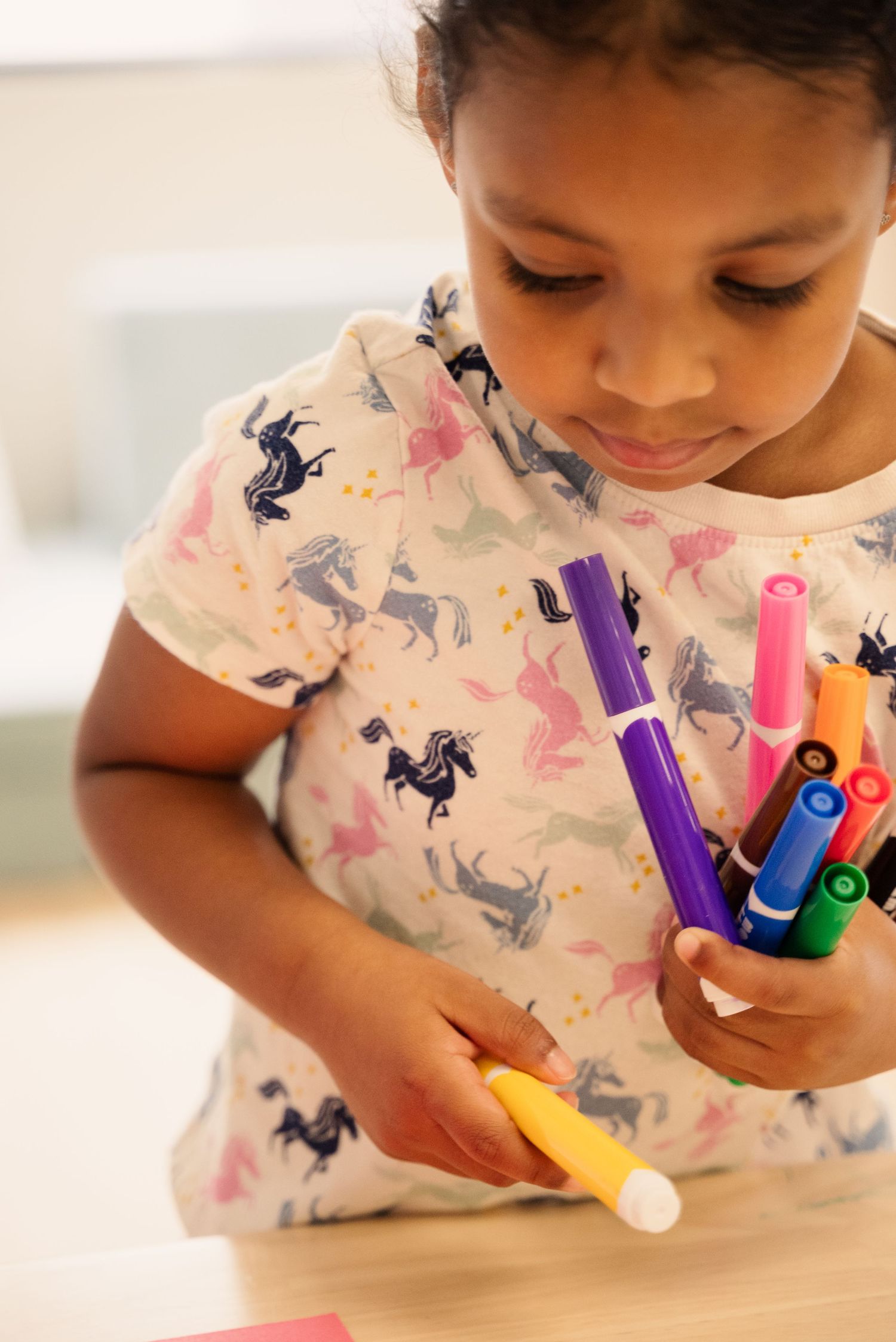 Child holding colorful markers while wearing a patterned shirt with unicorn designs.