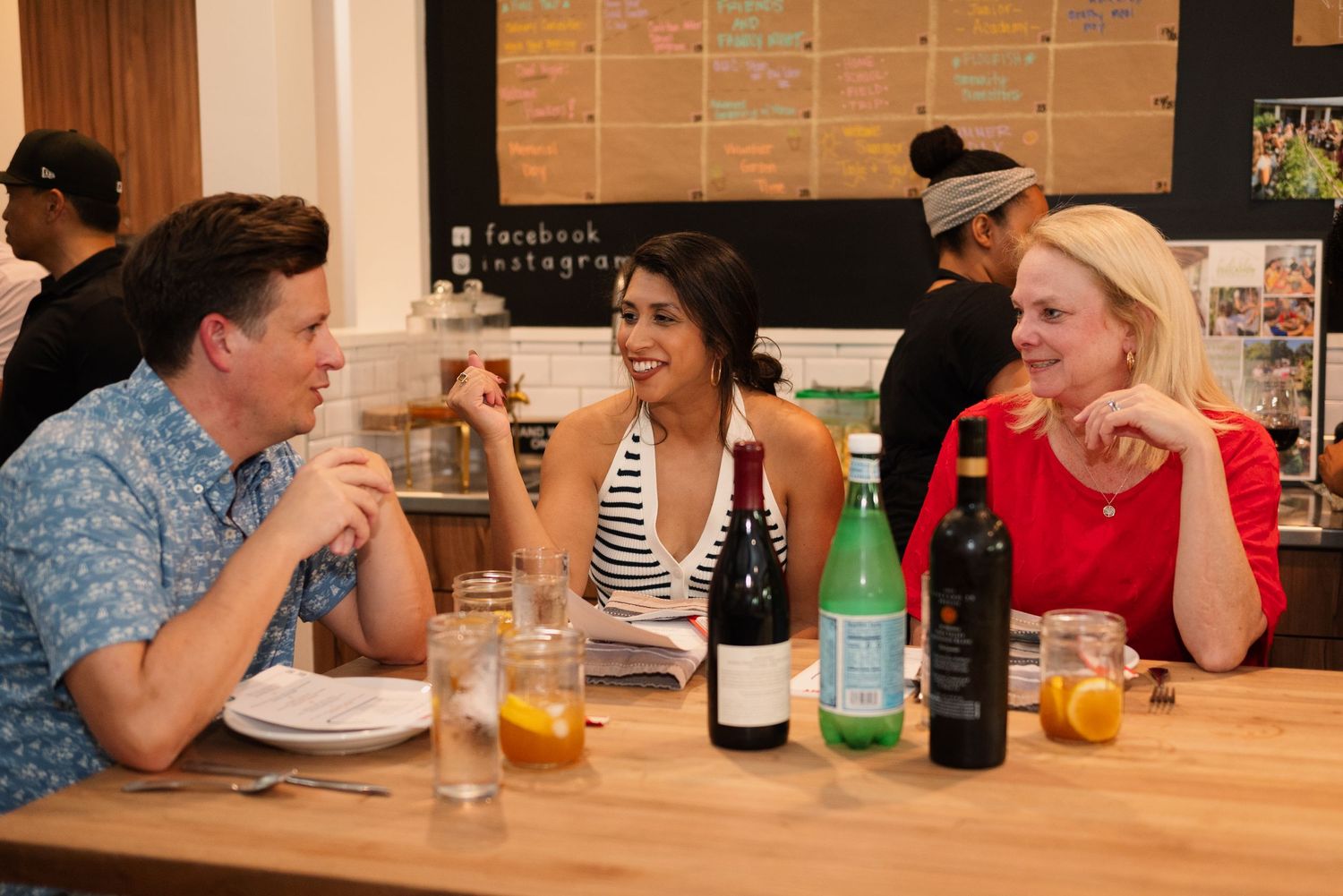 Group enjoying drinks and conversation at wooden table with wine bottles and beverages.