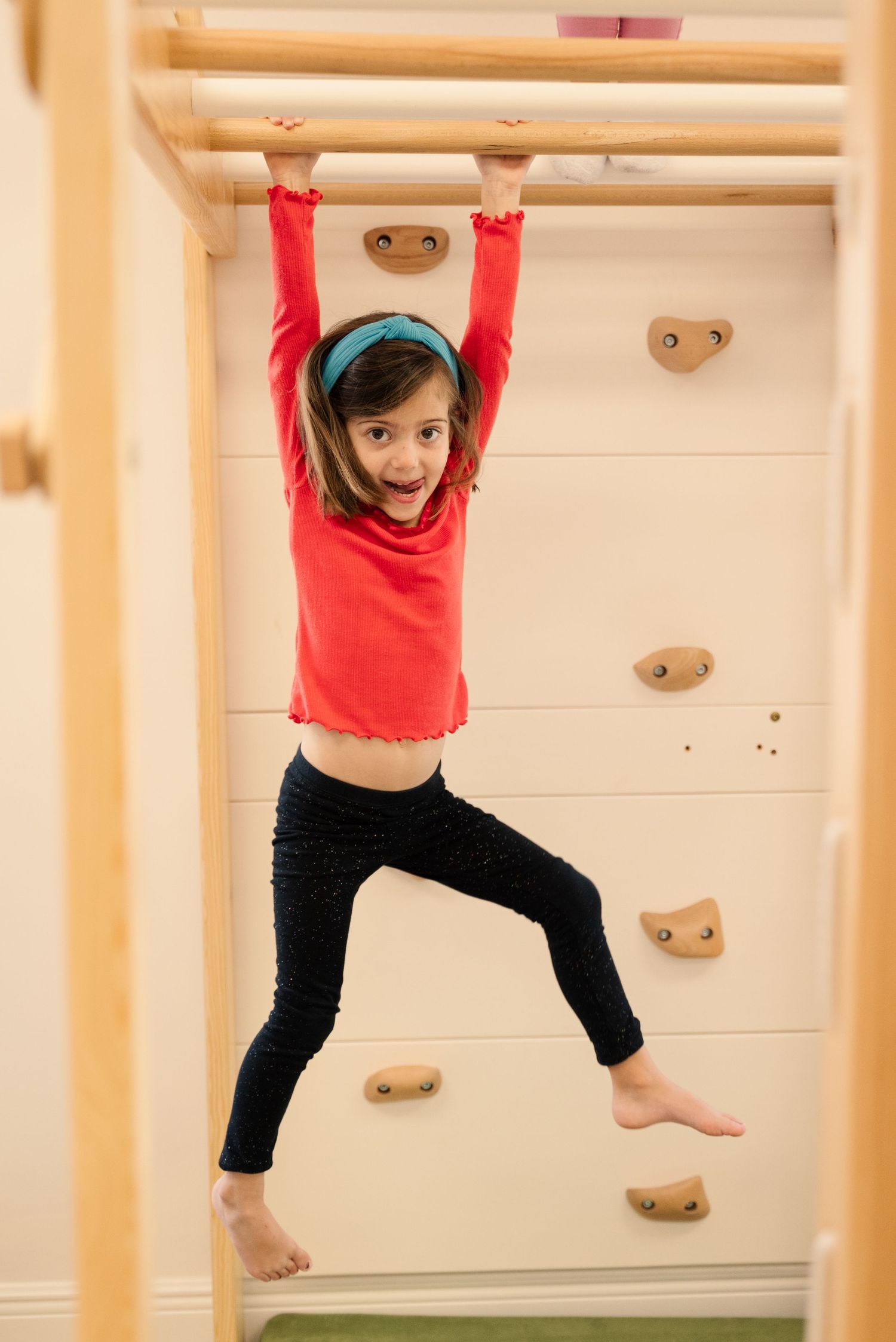 Child in red shirt and black pants hanging from wooden climbing bars.