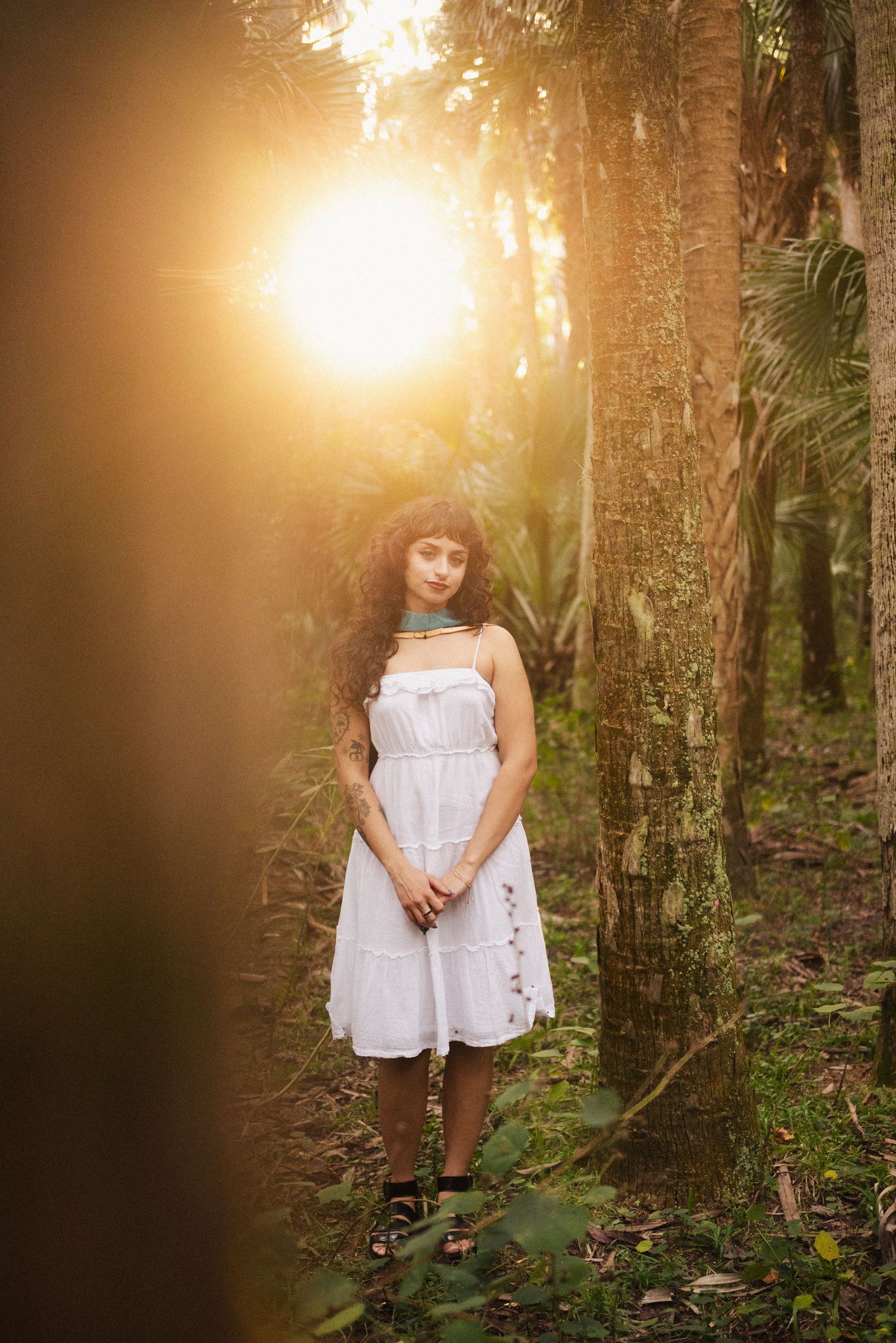 A white dress glows in golden sunlight filtering through mossy trees in a forest setting.