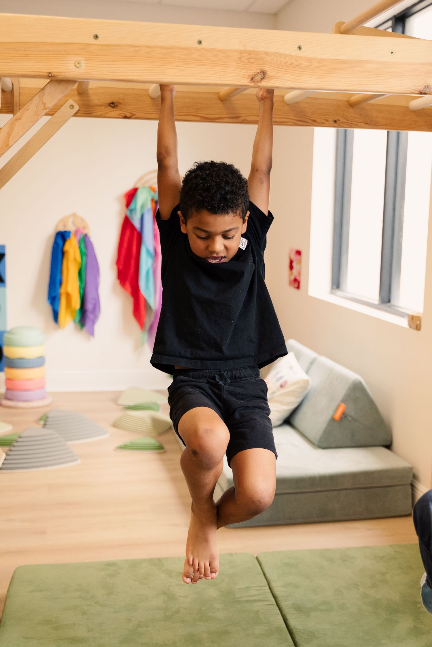 Child in black outfit climbing on wooden indoor play structure with green mat below.