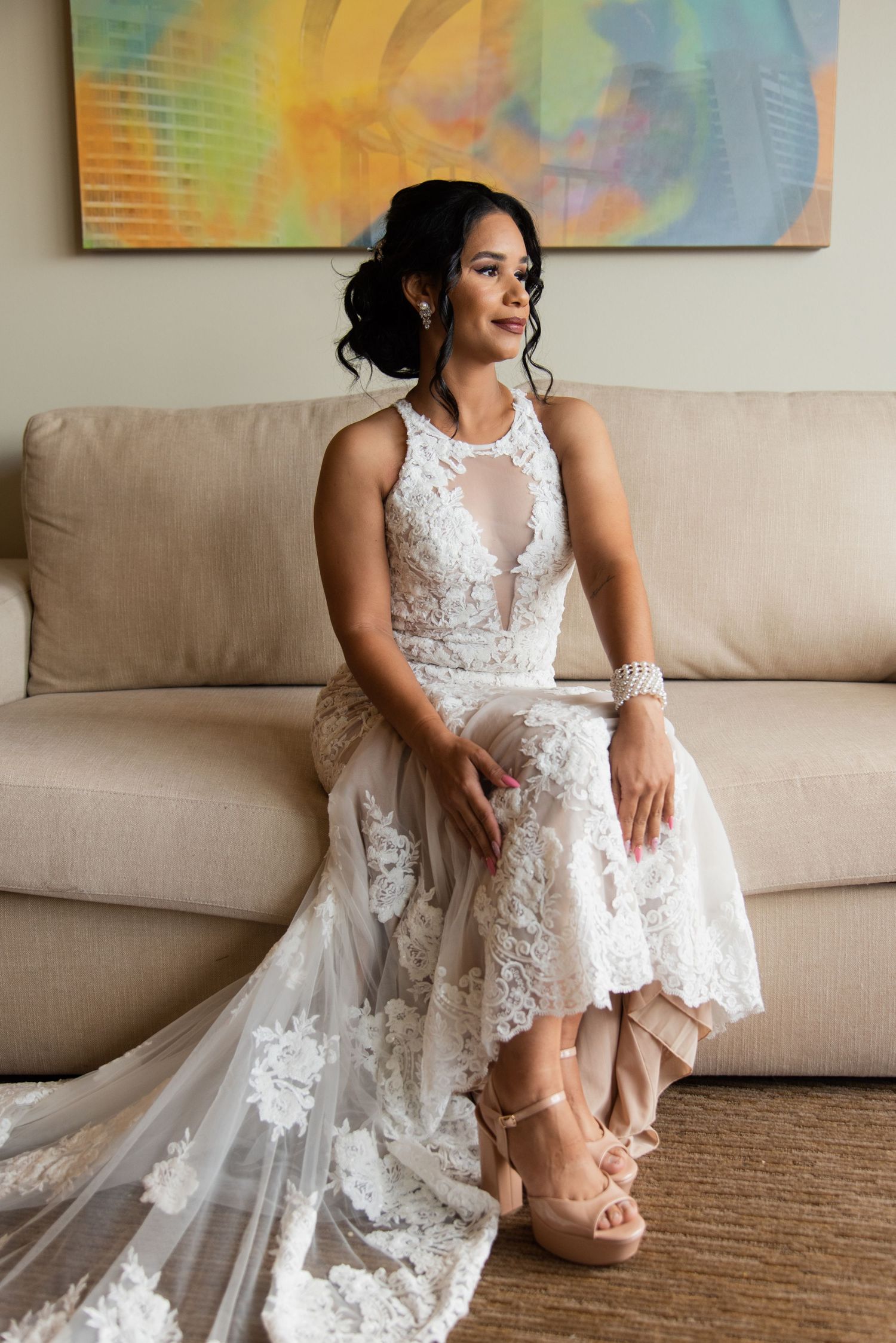Bride in detailed lace gown sits elegantly on beige couch with colorful artwork in background.
