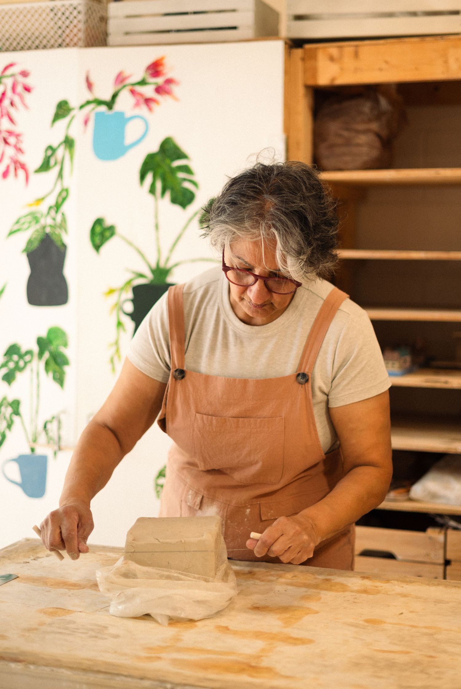 Potter working with clay at wooden table against wall decorated with tropical leaf motifs.
