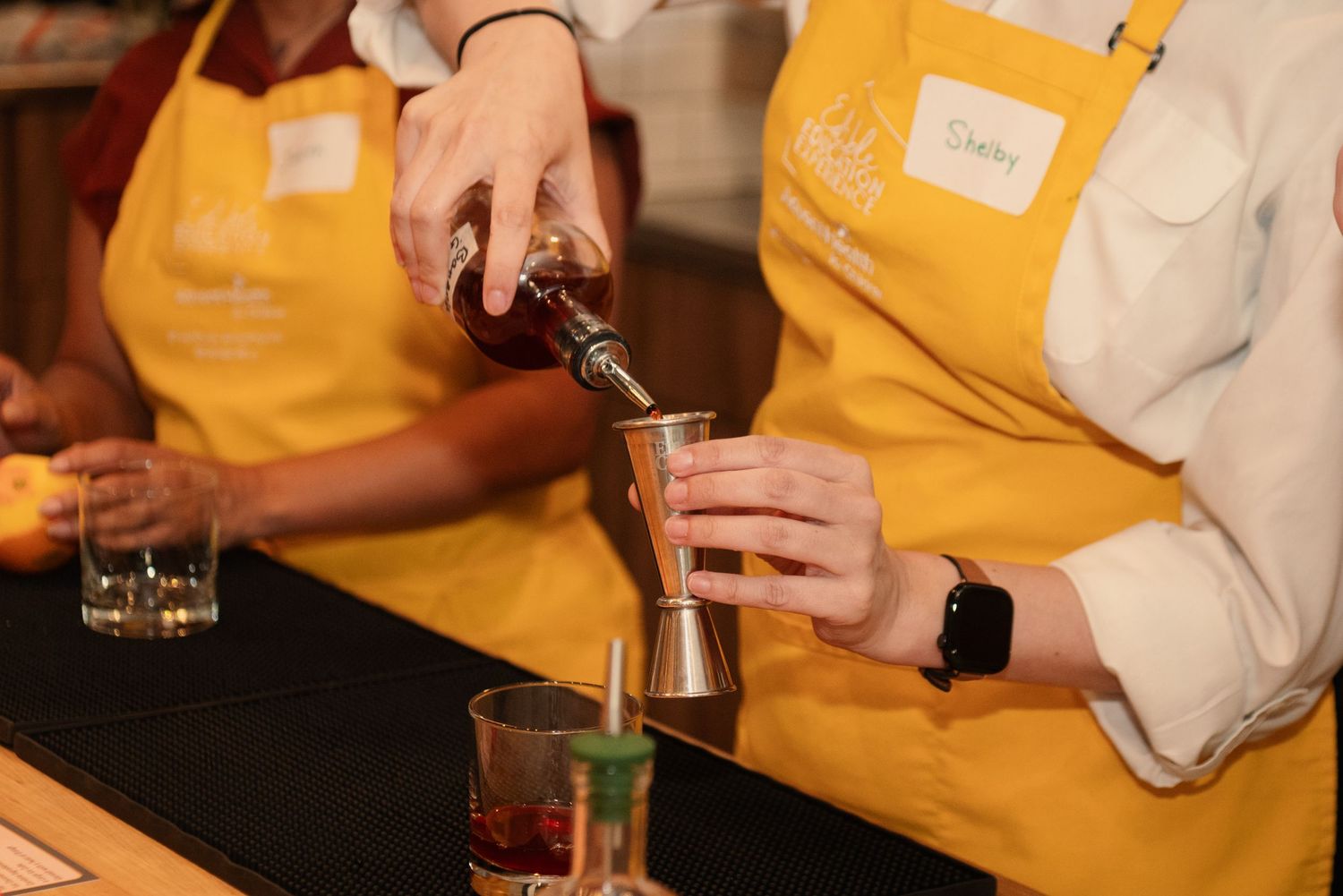 Close-up of hands pouring drinks during a cocktail making class with yellow aprons visible.