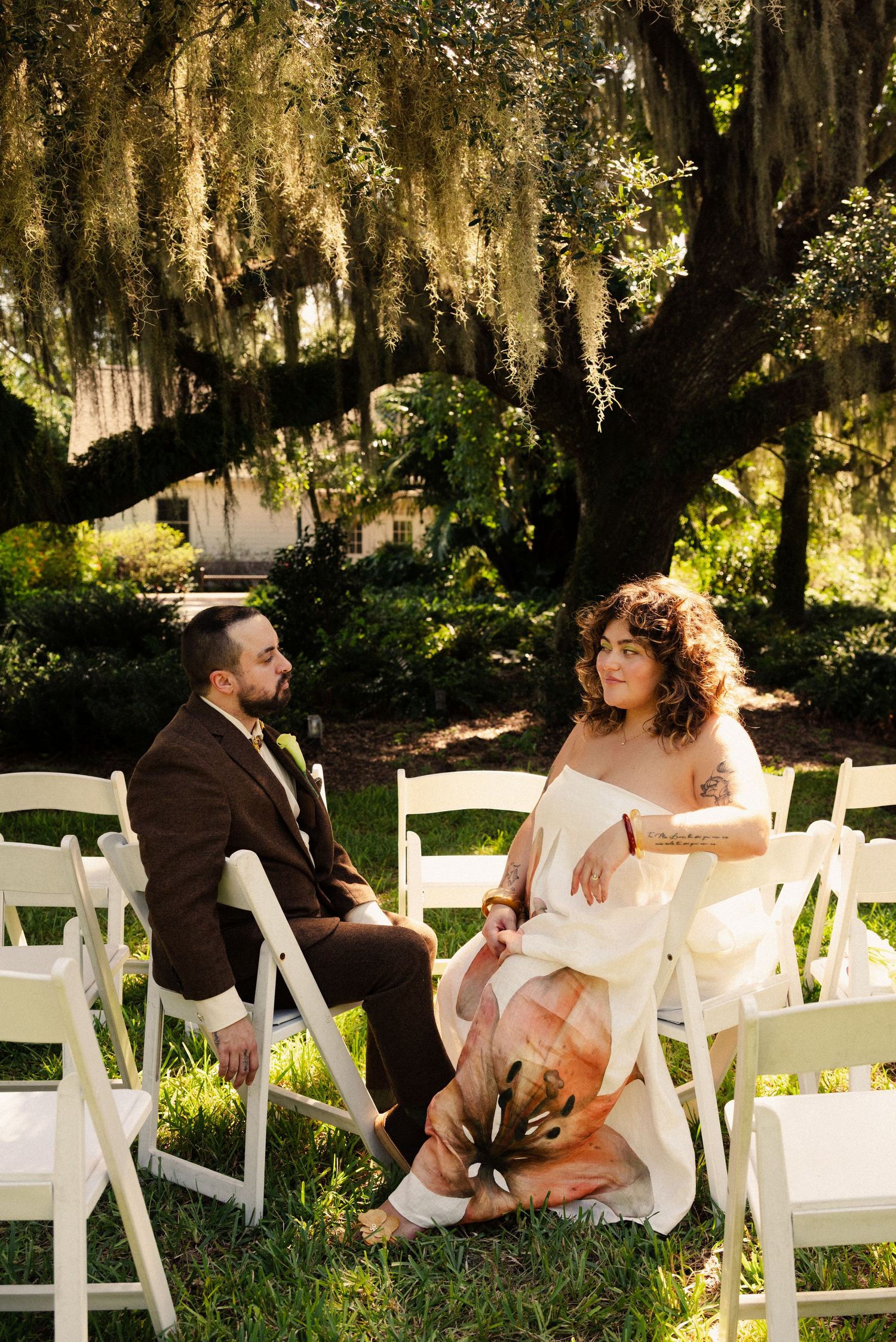 A couple sits together on white folding chairs under Spanish moss-draped trees.
