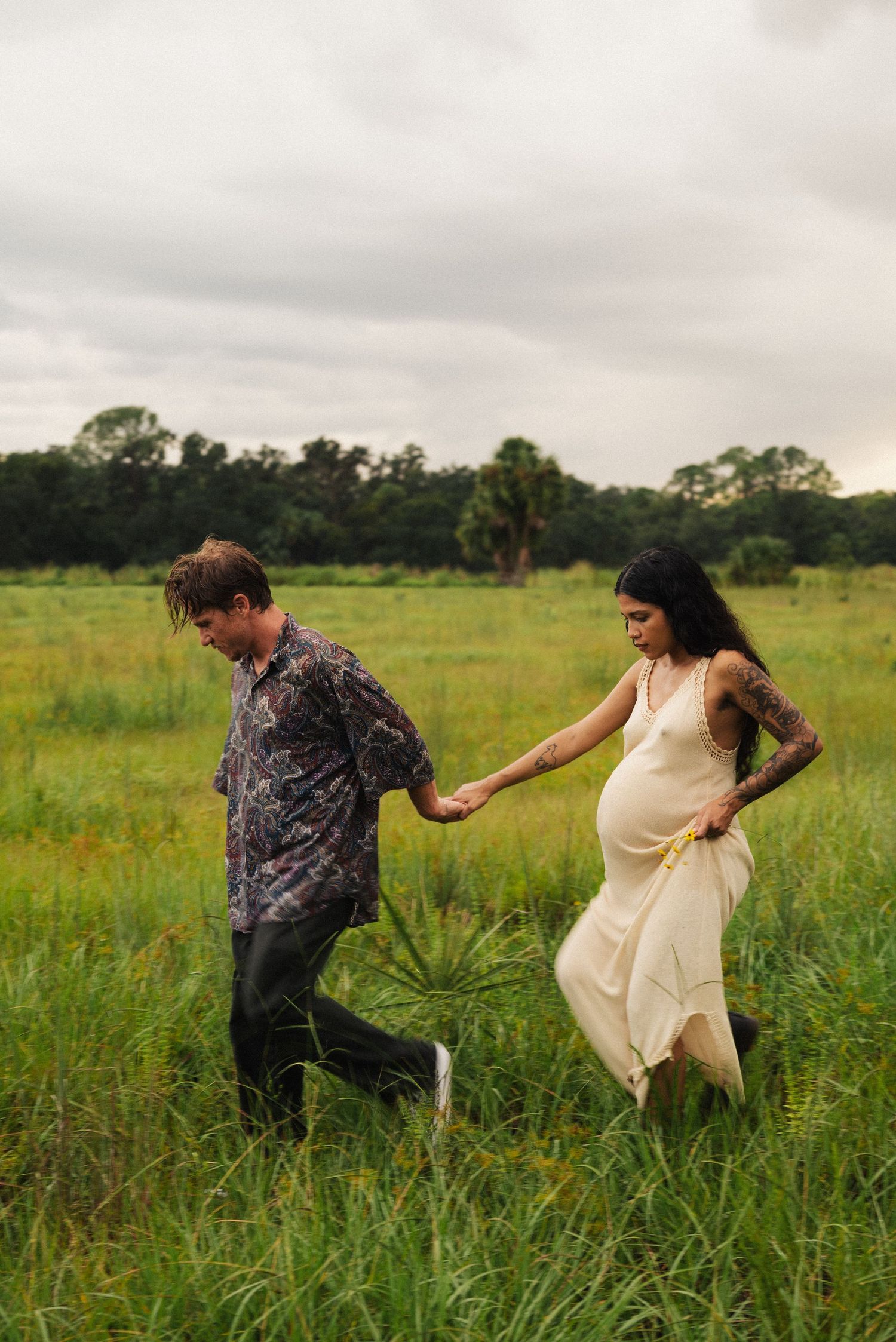 Couple running playfully through a green meadow on a cloudy day.