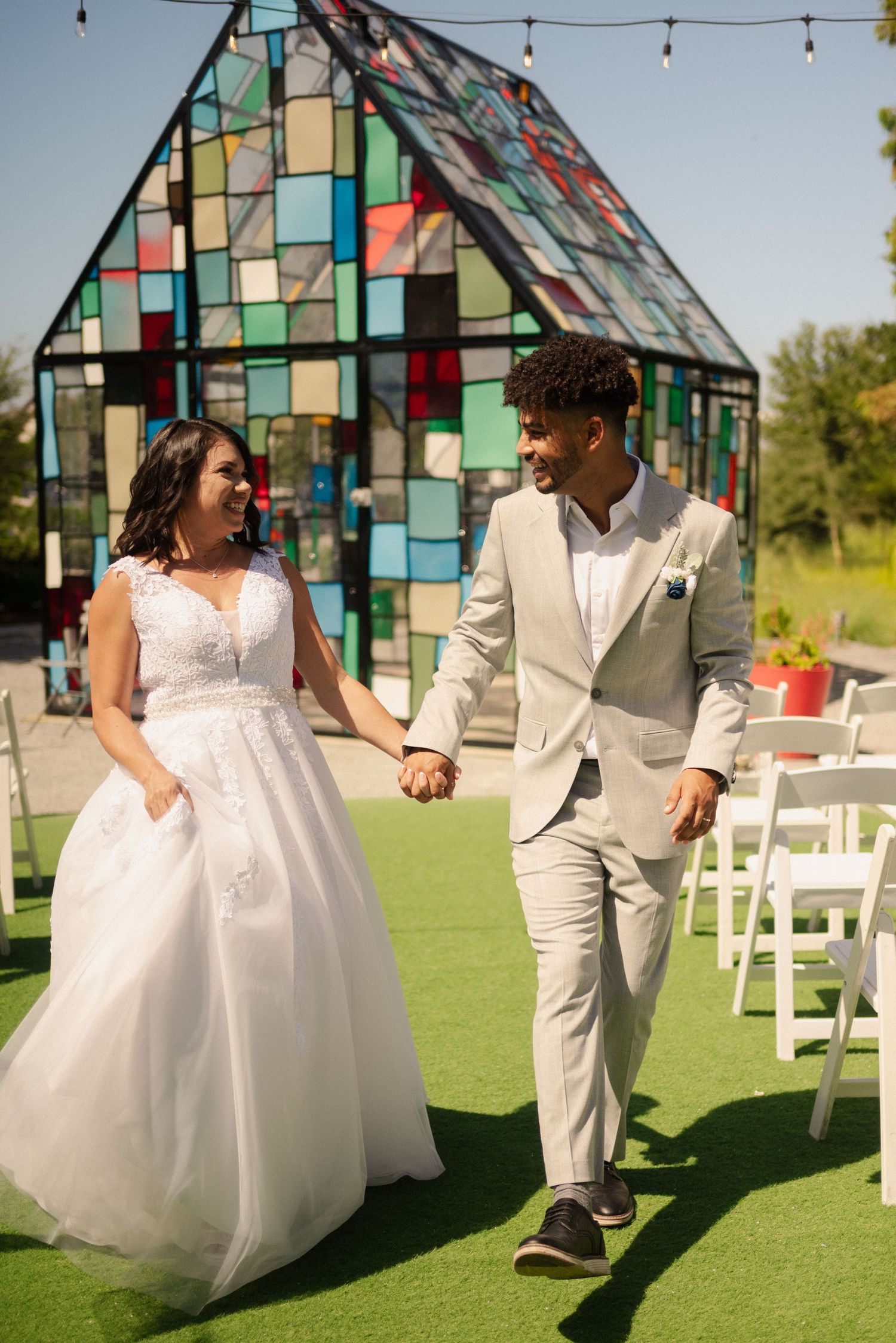 Couple dancing and laughing on green grass with a colorful stained glass chapel backdrop.