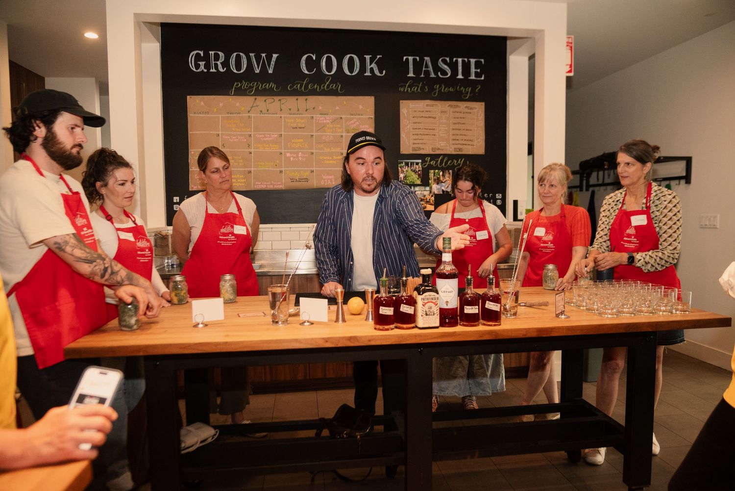 Students in red aprons learn cooking techniques under a 'Grow Cook Taste' sign in a modern kitchen.
