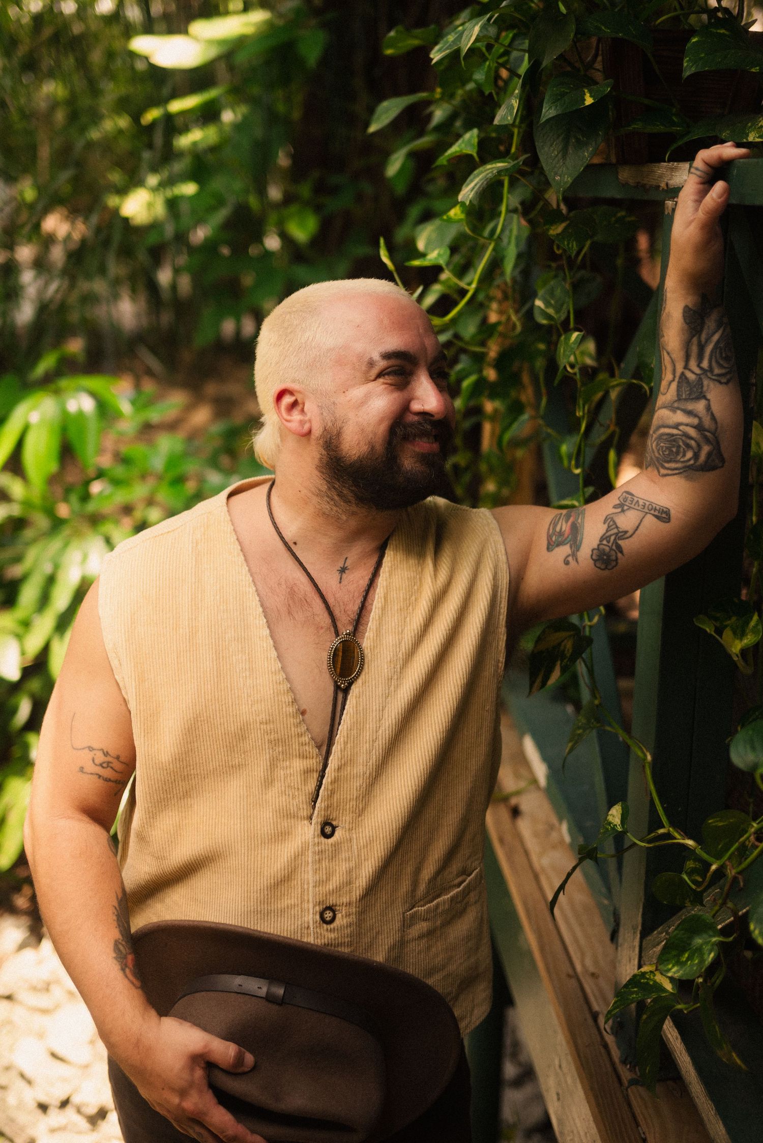 Person in tan vest poses thoughtfully against leafy backdrop in warm sunlight.